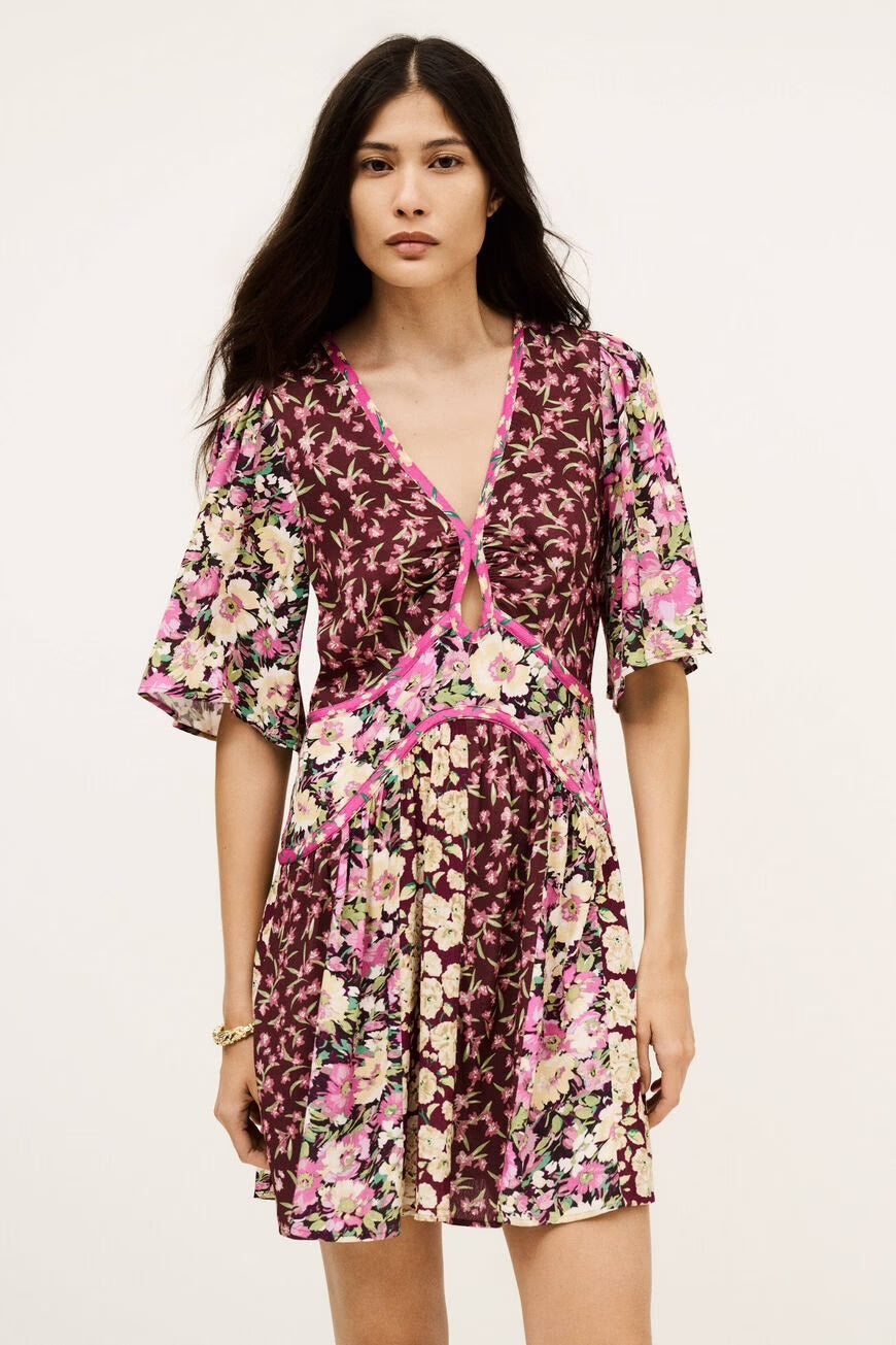 Frau trägt ein kurzärmeliges, kurz geschnittenes Kleid mit floralem Muster in Rosa, Creme, Bordeaux und Grün. Das Kleid hat einen V-Ausschnitt mit kleinem Cut-out unter der Brust und eine figurbetonte Taille mit Ziernähten in Pink. Der Rock ist leicht ausgestellt und besteht wie das Oberteil aus unterschiedlichen geblümten Stoffbahnen.