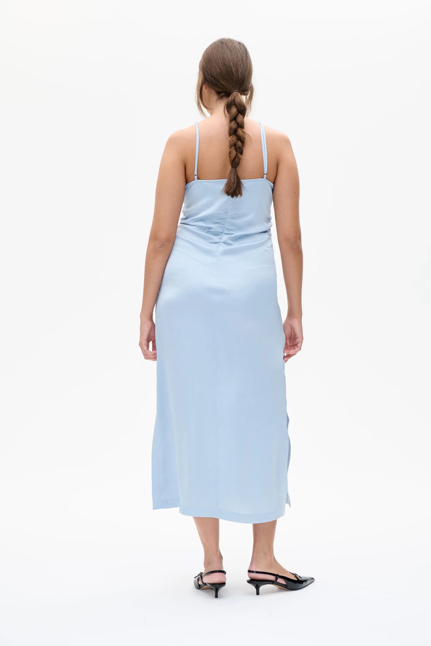 Frau trägt ein hellblaues, ärmelloses Midikleid mit Spaghettiträgern und V-Ausschnitt. Das Kleid hat eine geraffte Partie am Rücken und fällt figurbetont bis midiwadenlang. Sie kombiniert es mit schwarzen Slingback-Pumps mit kleinem Absatz. Der Hintergrund ist weiß und neutral gehalten.