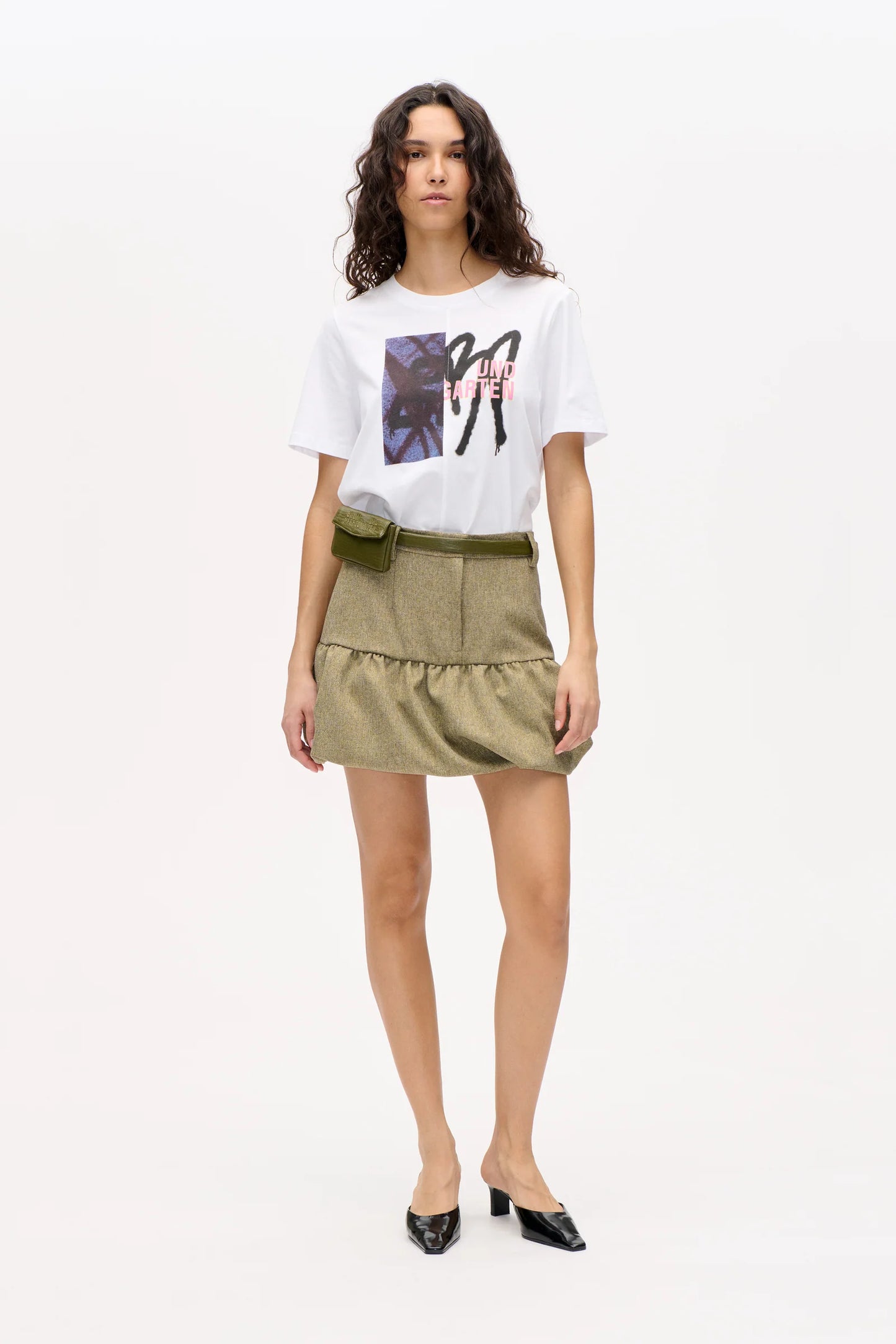 Frau vor weißer Wand, sie trägt einen grünen Tweed-Rock und weißes Printshirt.