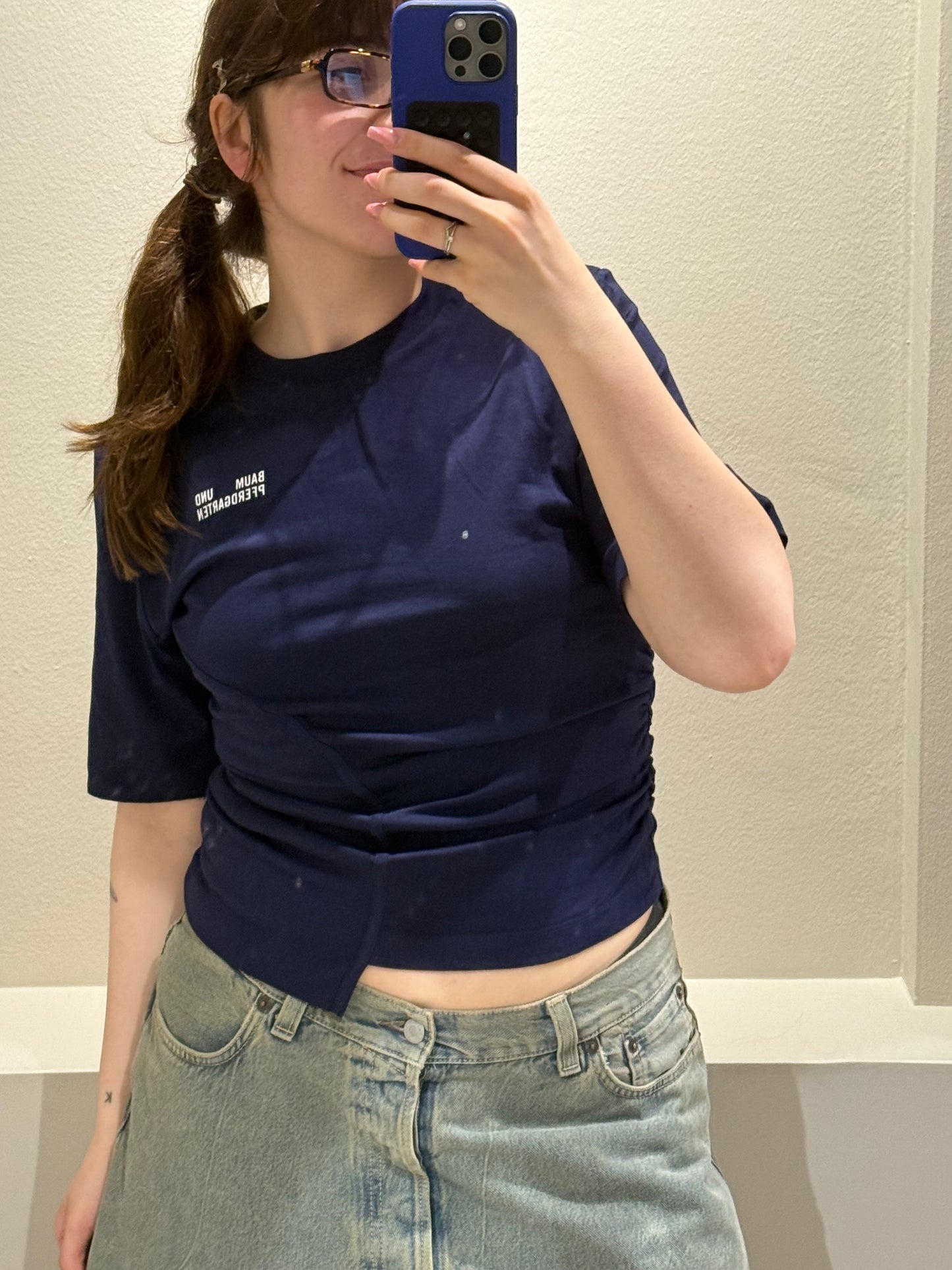 Frau trägt ein dunkelblaues, tailliertes T-Shirt mit asymmetrischem Schnitt und seitlicher Raffung. Auf der linken Brust befindet sich eine kleine Logo-Aufschrift ‚Baum und Pferdgarten‘. Dazu kombiniert sie eine hellblaue Jeans mit weitem Bein. Sie steht in einer Umkleidekabine mit hellem Vorhang und violettem Hocker.