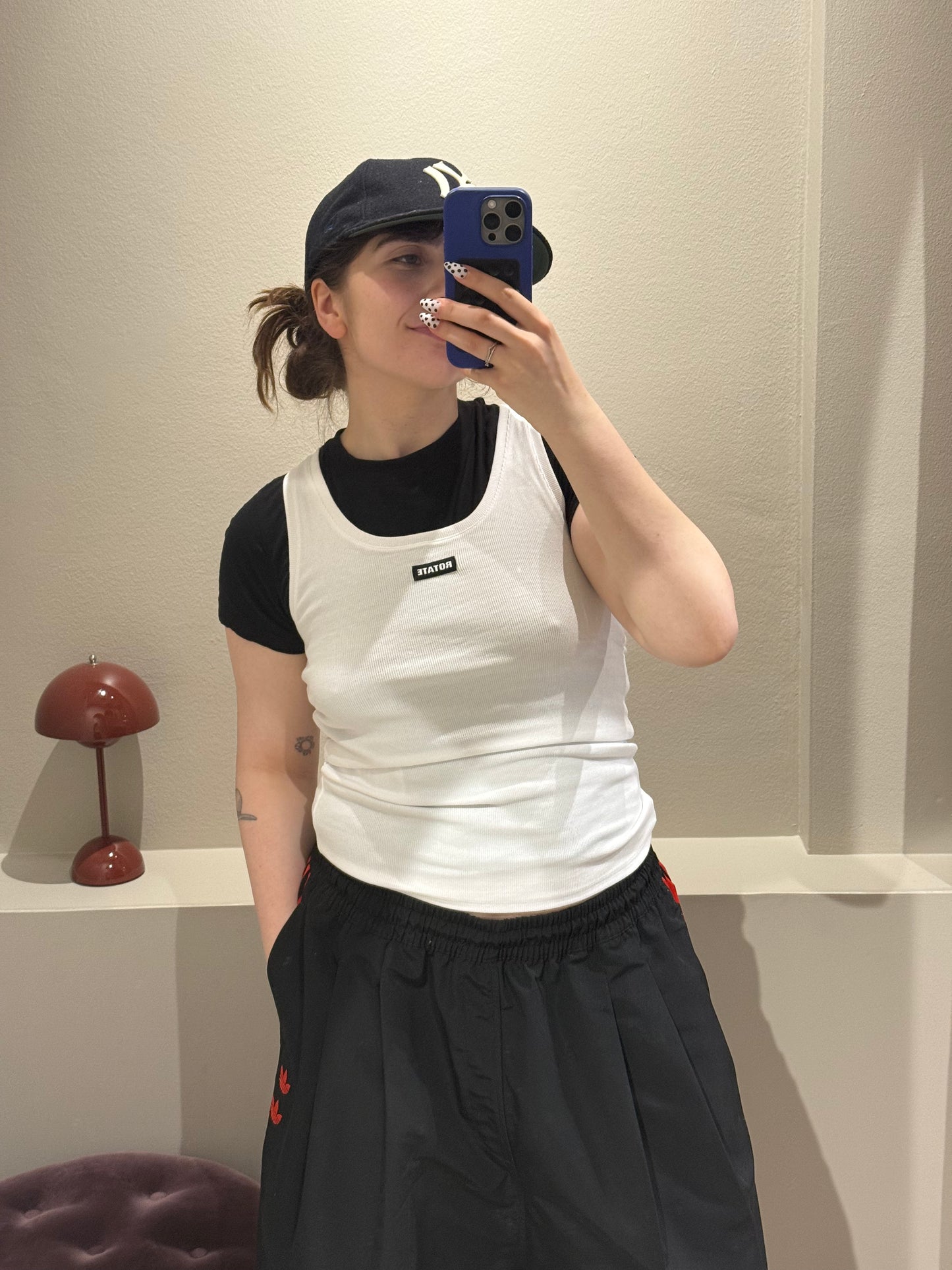 Frau macht Spiegelselfie, sie trägt ein weißes Tanktop über einem schwarzem Tshirt. Das Top hat einen U-Ausschnitt und ein Rotate Logopatch mittig auf der Brust. Dazu Kombiniert trägt sie eine schwarze Hose.