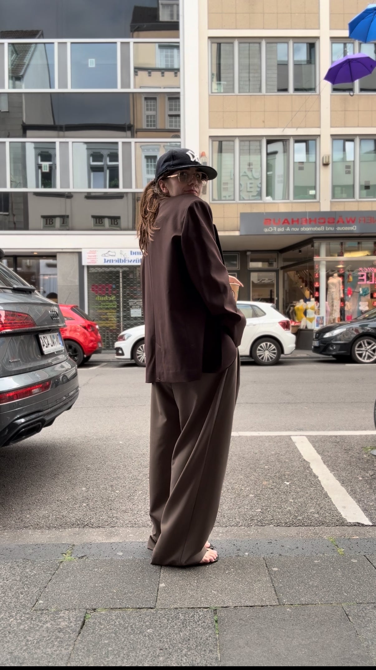 Frau trinkt Kaffe, sie trägt eine braune Hose mit einem overiszed Blazer, der ist schokobraun. Im Hintergrund sieht man eine Straße. Diesmal im Seitenprofil.