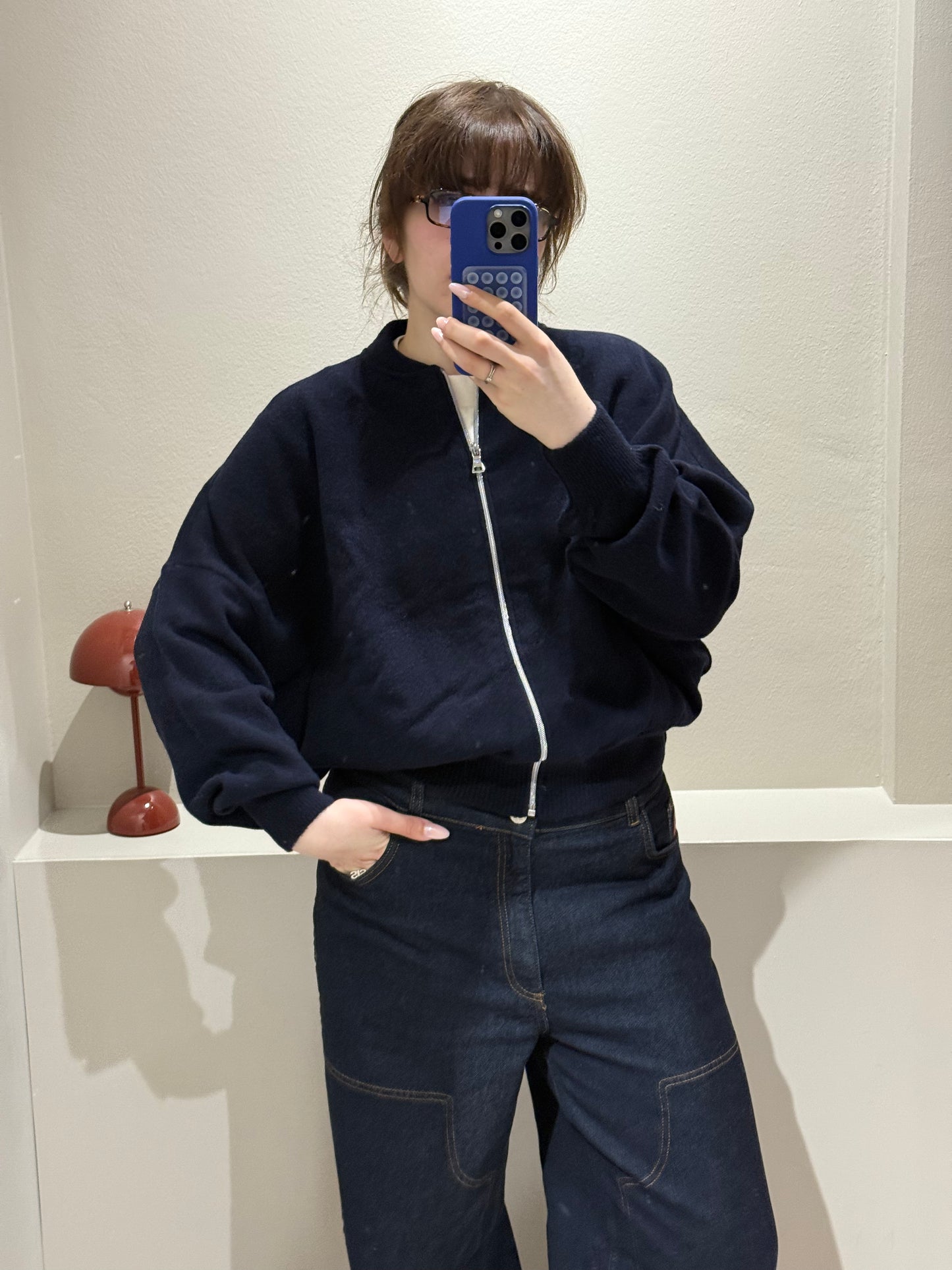 Frau macht Spiegelselfie, sie trägt iene dunkelblaue Strickbomberjacke und dunkle Jeans.
