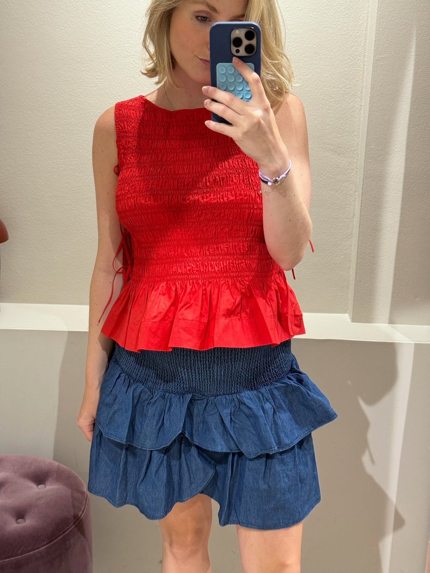 Frau steht in einer Umkleidekabine und macht ein Spiegelselfie. Sie trägt ein ärmelloses, rotes Top mit gesmoktem Oberteil und Volant am Saum. Dazu kombiniert sie einen dunkelblauen Minirock mit gesmoktem Bund und doppeltem Volant aus Jeansstoff. Ihr blondes Haar ist offen, und sie hält das Handy vor ihr Gesicht.