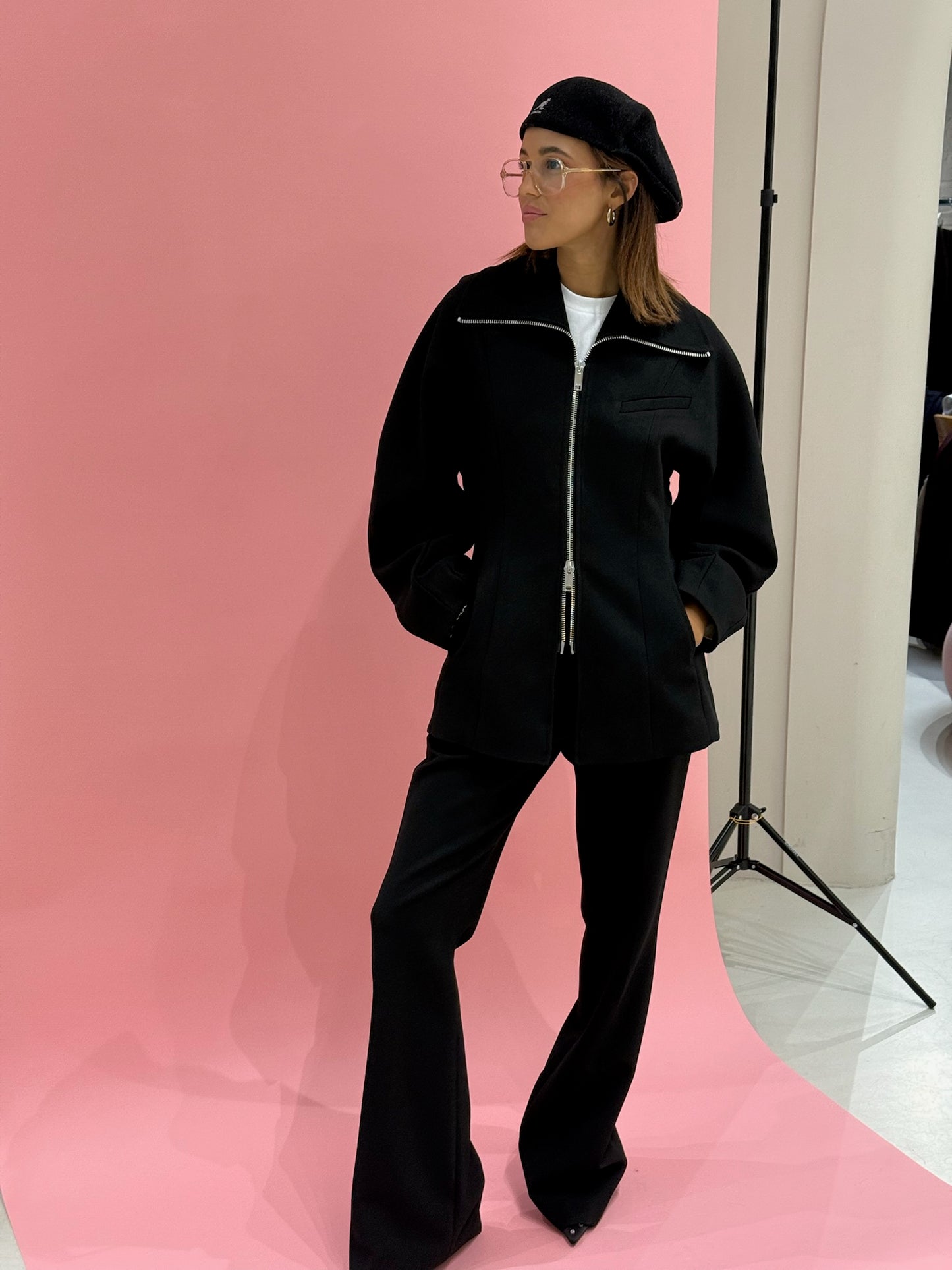 Frau vor pinken Hintergrund, sie trägt schwarzen Blazer und eine schwarze Hose, dazu eine schwarze Schirmmütze.