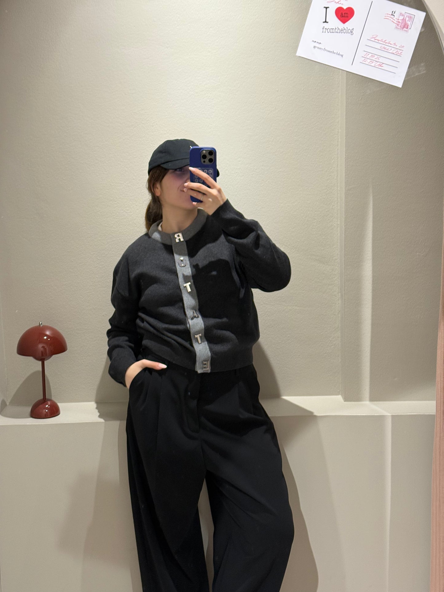 Frau macht Spiegelselfie, sie trägt eine schwarze Hose und einen grauen Cardigan mit Kontrastsäumen und Rotate geschrieben an der Knopfleiste.