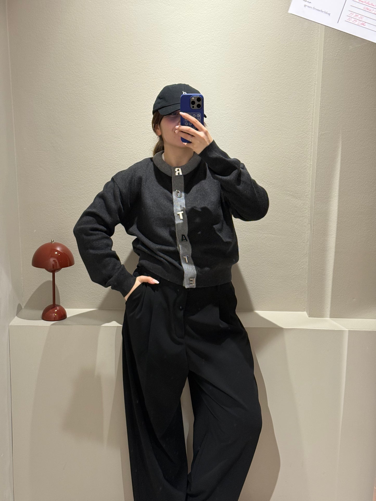 Frau macht Spiegelselfie, sie trägt eine schwarze Hose und einen grauen Cardigan mit Kontrastsäumen und Rotate geschrieben an der Knopfleiste.