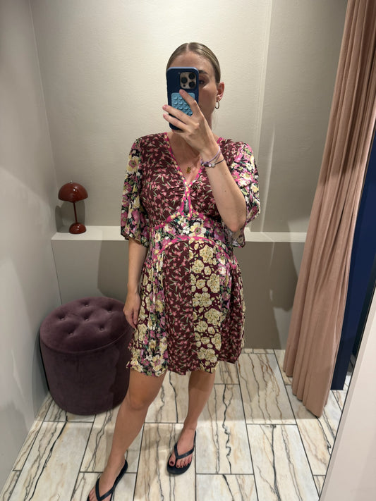 Frau steht in einer Umkleidekabine und macht ein Spiegelselfie. Sie trägt ein geblümtes, bordeauxfarbenes Kleid mit V-Ausschnitt, kurzem Schnitt und halblangen Ärmeln. Das Kleid hat ein Patchwork-Muster mit floralen Prints in Rosa, Creme und Grün sowie pinkfarbene Zierpaspeln. Der leicht ausgestellte Rock fällt locker über einen sichtbaren Babybauch. Die Frau trägt schwarze Zehensandalen und hat ihre blonden Haare streng zurückgebunden