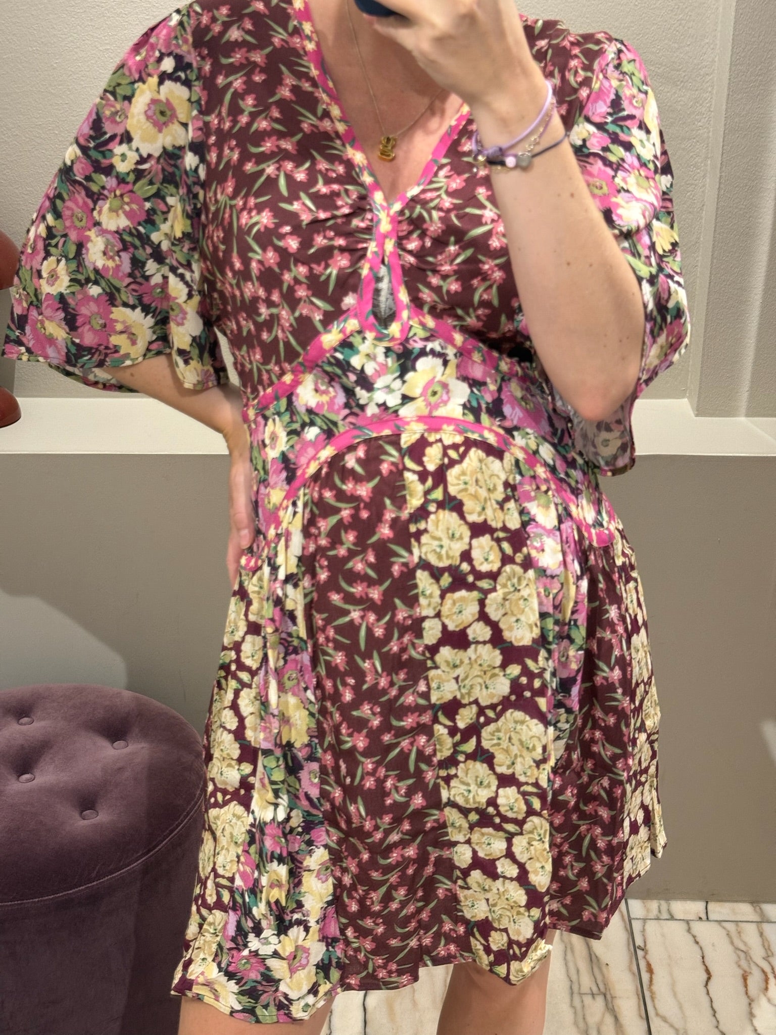 Frau steht in einer Umkleidekabine und macht ein Spiegelselfie. Sie trägt ein geblümtes, bordeauxfarbenes Kleid mit V-Ausschnitt, kurzem Schnitt und halblangen Ärmeln. Das Kleid hat ein Patchwork-Muster mit floralen Prints in Rosa, Creme und Grün sowie pinkfarbene Zierpaspeln. Der leicht ausgestellte Rock fällt locker über einen sichtbaren Babybauch. Die Frau trägt schwarze Zehensandalen und hat ihre blonden Haare streng zurückgebunden.