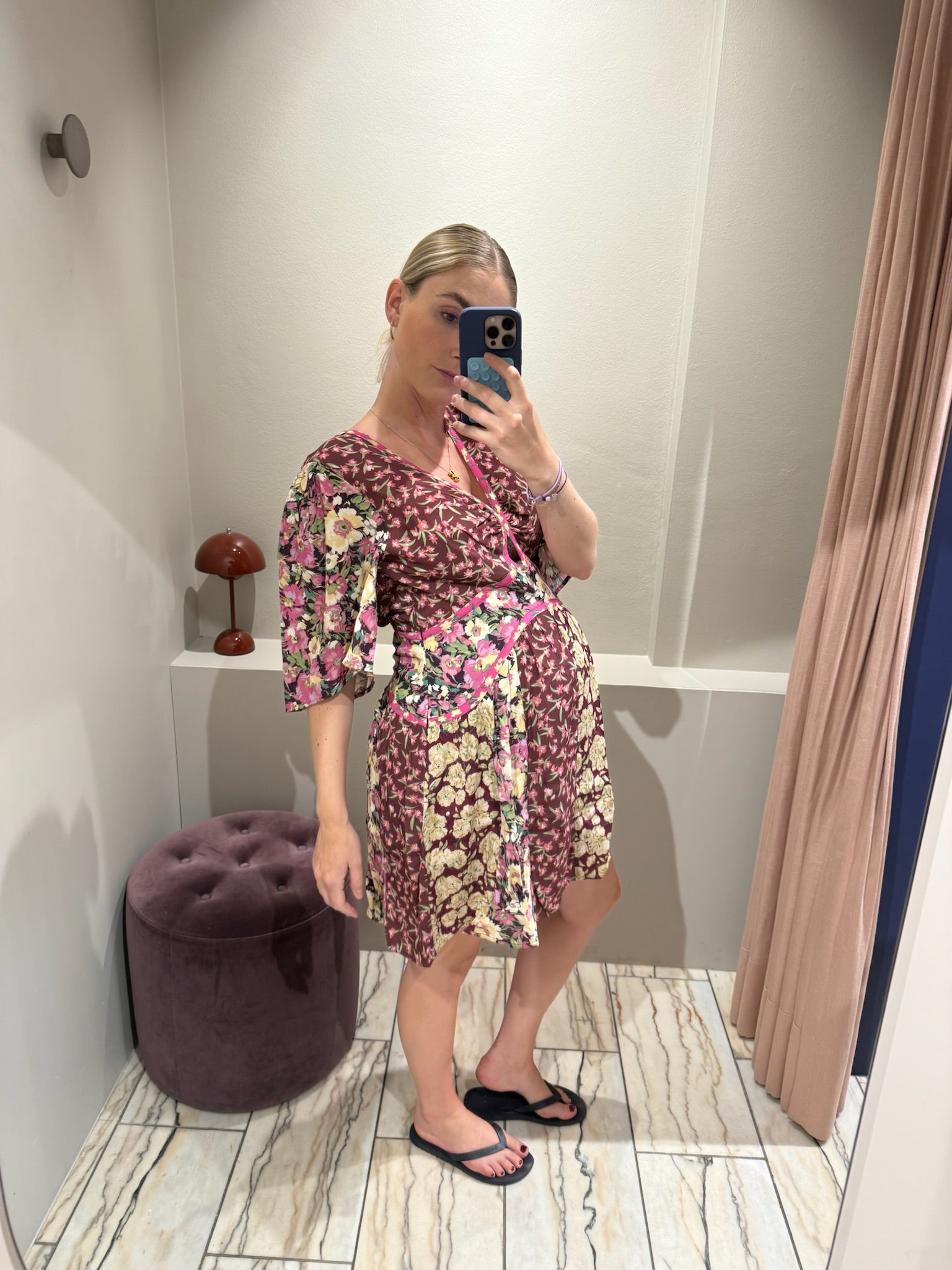 Frau steht in einer Umkleidekabine und macht ein Spiegelselfie. Sie trägt ein geblümtes, bordeauxfarbenes Kleid mit V-Ausschnitt, kurzem Schnitt und halblangen Ärmeln. Das Kleid hat ein Patchwork-Muster mit floralen Prints in Rosa, Creme und Grün sowie pinkfarbene Zierpaspeln. Der leicht ausgestellte Rock fällt locker über einen sichtbaren Babybauch. Die Frau trägt schwarze Zehensandalen und hat ihre blonden Haare streng zurückgebunden.