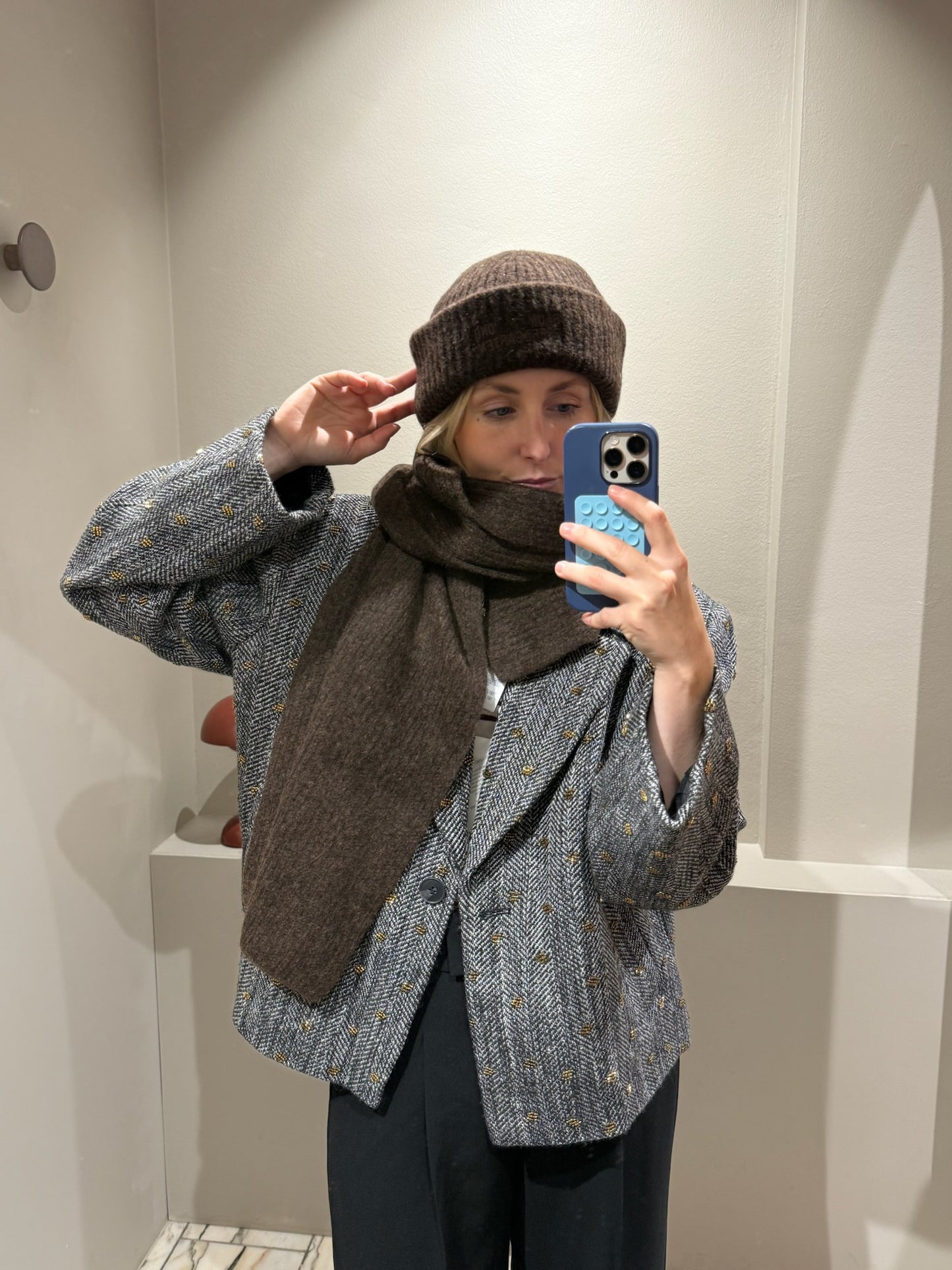 Frau macht Spiegelselfie, sie trägt eine schwarze Hose dazu einen grauen Blazer mit Pailletten und braune Mütze und Schal.