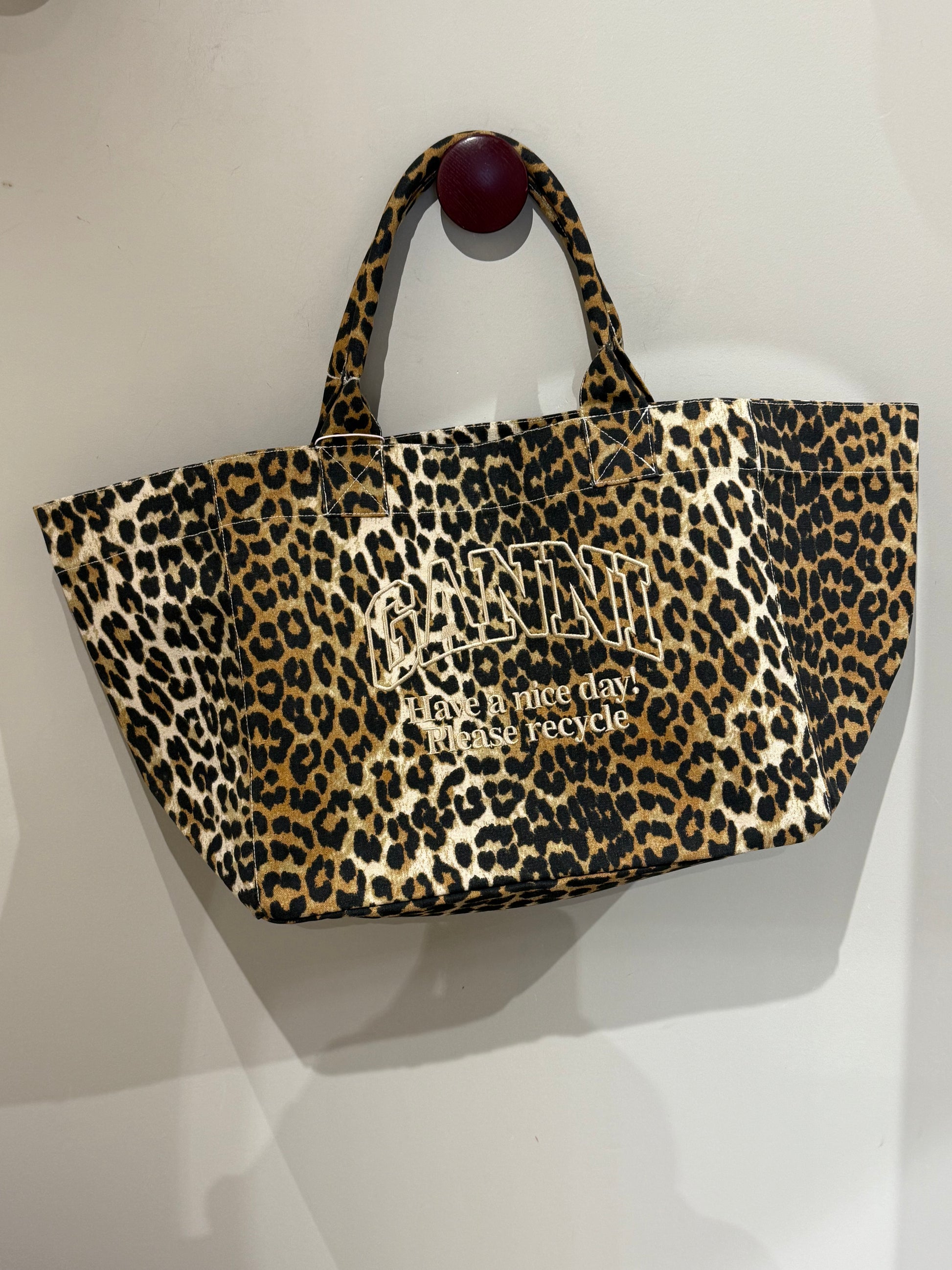 XXL-Shopper von Ganni aus recyceltem Baumwoll-Canvas mit Leopardenmuster, aufgesticktem „Have a nice day! Please recycle“-Schriftzug, zwei stabilen Tragegriffen und offenem Innenfach. Große, lässige Form.
