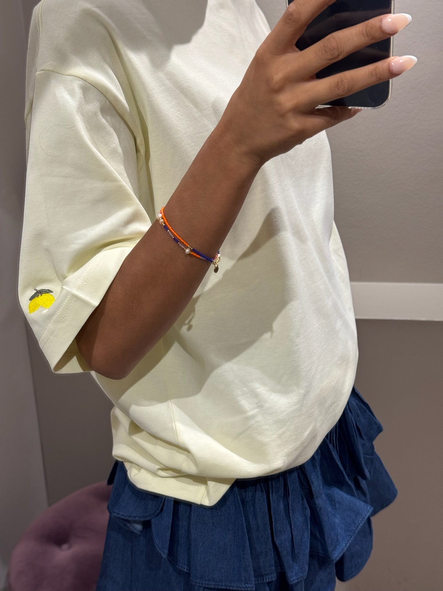 ine Frau steht in einer Umkleidekabine und macht ein Selfie vor einem Spiegel. Sie trägt ein oversized T-Shirt in zartem Buttergelb mit kurzen Ärmeln. Auf der linken Brust ist ein gesticktes ‚G‘ zu sehen, am linken Ärmel eine kleine Zitronen-Stickerei. Dazu kombiniert sie einen kurzen, dunkelblauen Stufenrock mit gesmoktem Bund und Volants. Ihr Gesicht ist größtenteils vom Smartphone verdeckt
