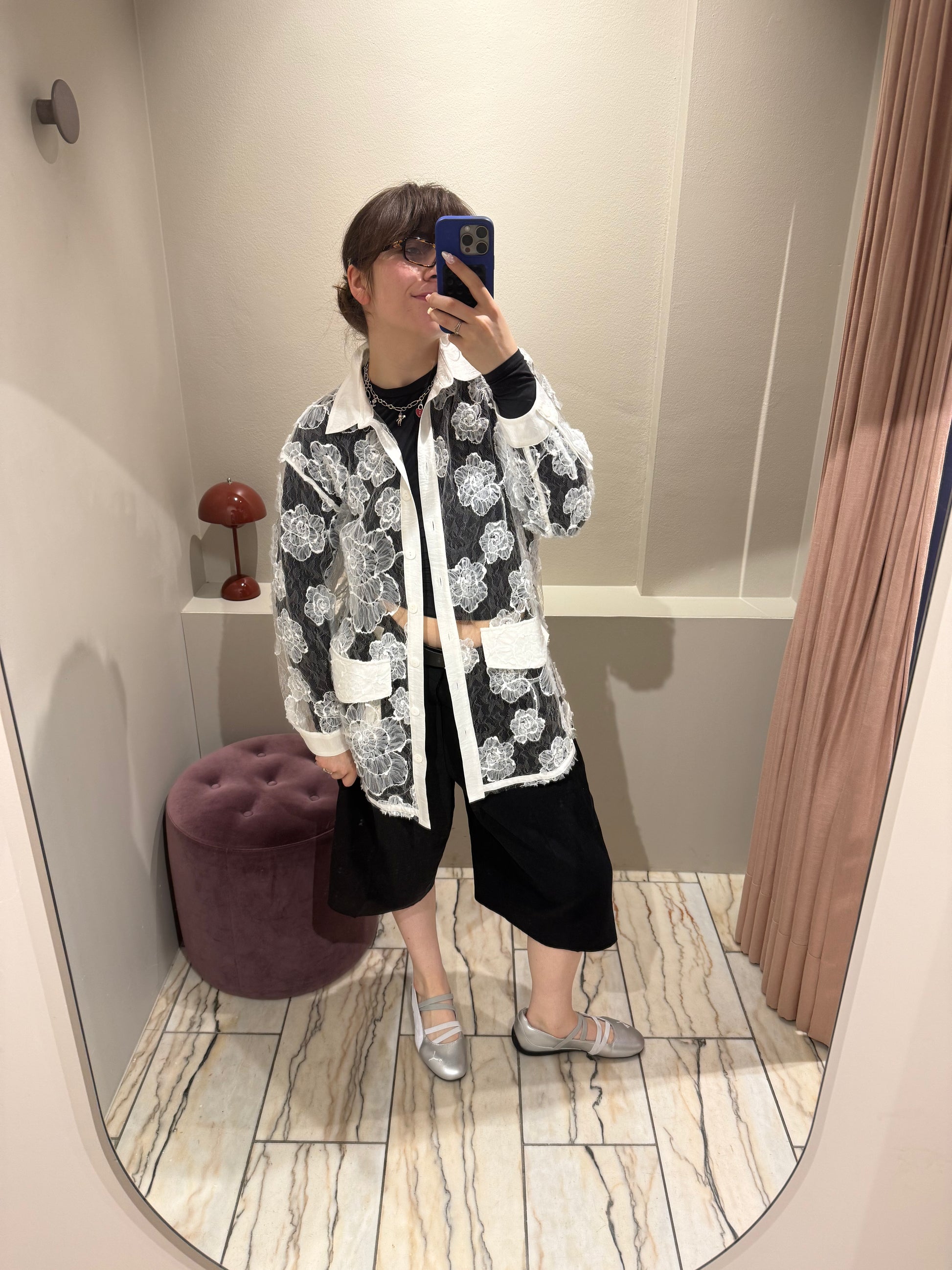 Eine Person steht in einer Umkleidekabine und macht ein Spiegel-Selfie. Sie trägt eine lange, transparente Bluse aus weißer Spitze mit großflächigem Blumenmuster. Die Bluse hat einen weißen Kragen, Knopfleiste, Manschetten und aufgesetzte Taschen. Darunter ist dunkle Kleidung sichtbar, die durch den transparenten Stoff hindurchscheint.