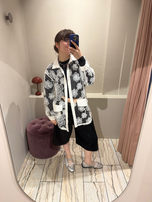 Eine Person steht in einer Umkleidekabine und macht ein Spiegel-Selfie. Sie trägt eine lange, transparente Bluse aus weißer Spitze mit großflächigem Blumenmuster. Die Bluse hat einen weißen Kragen, Knopfleiste, Manschetten und aufgesetzte Taschen. Darunter ist dunkle Kleidung sichtbar, die durch den transparenten Stoff hindurchscheint.