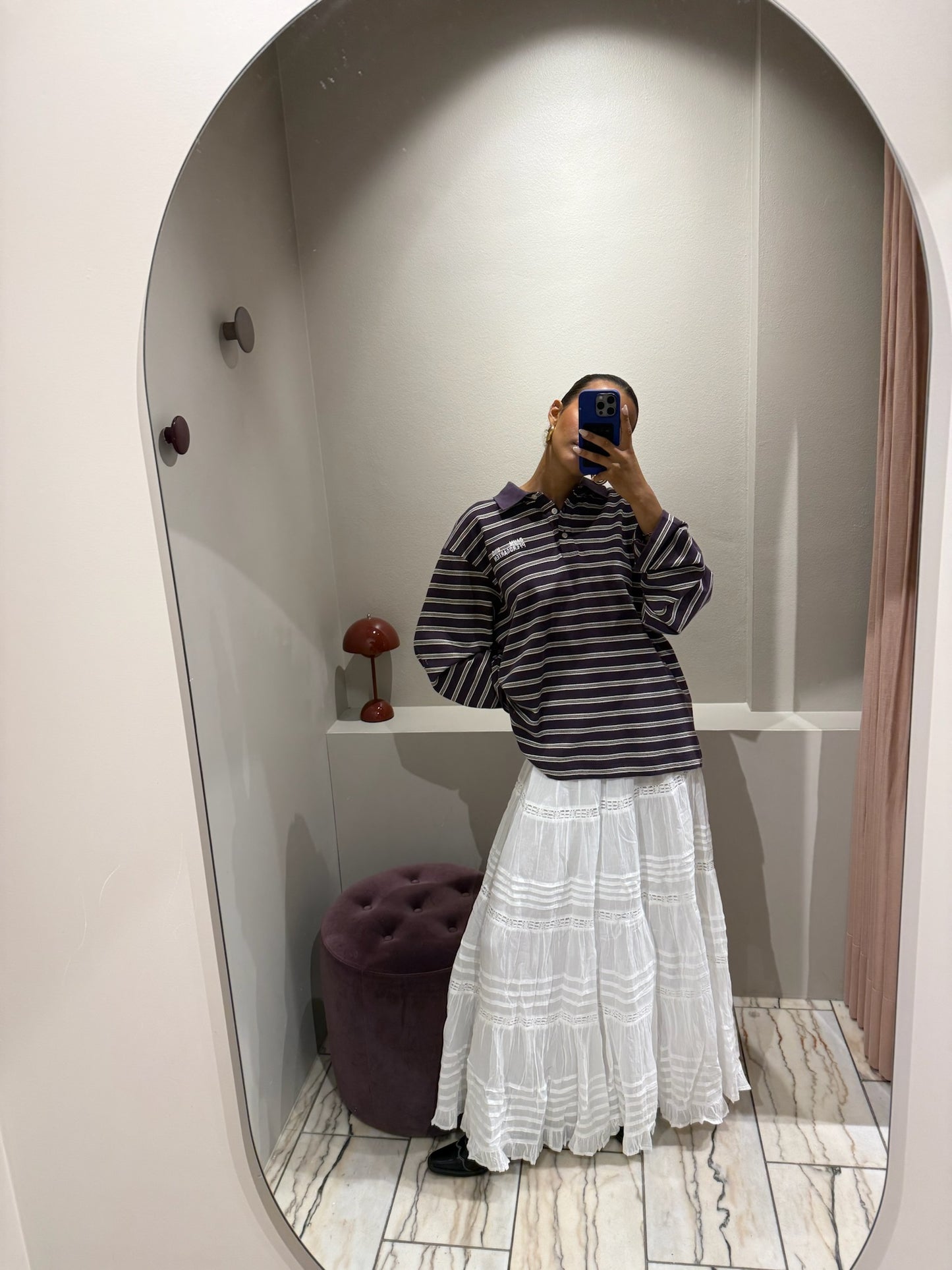 Eine Frau macht ein Spiegel Selfie, sie trägt einen weißen Rock mit horizontalen Lochstick-Streifen und Rüschensaum. Der rock ist sehr romantisch und fällt locker. Dazu styled sie ein oversized Polo und Cowboyboots.