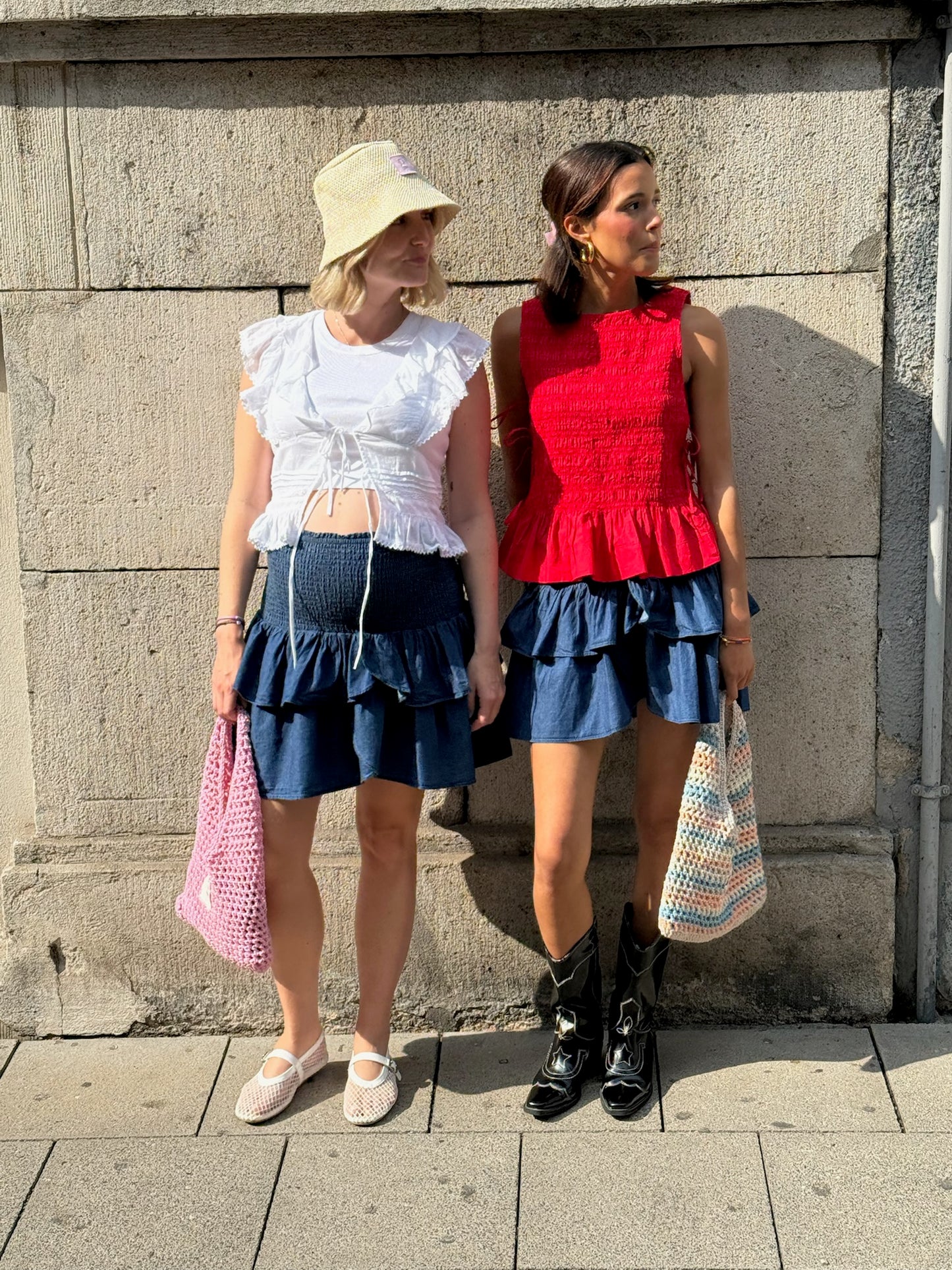 Zwei Frauen stehen nebeneinander vor einer Steinwand in der Sonne. Beide tragen blaue Stufenröcke mit Volants. Die eine kombiniert ihn mit weißem Volant-Top, Sonnenhut und Ballerinas, die andere mit rotem Top, Cowboy-Boots und gehäkelter Tasche.