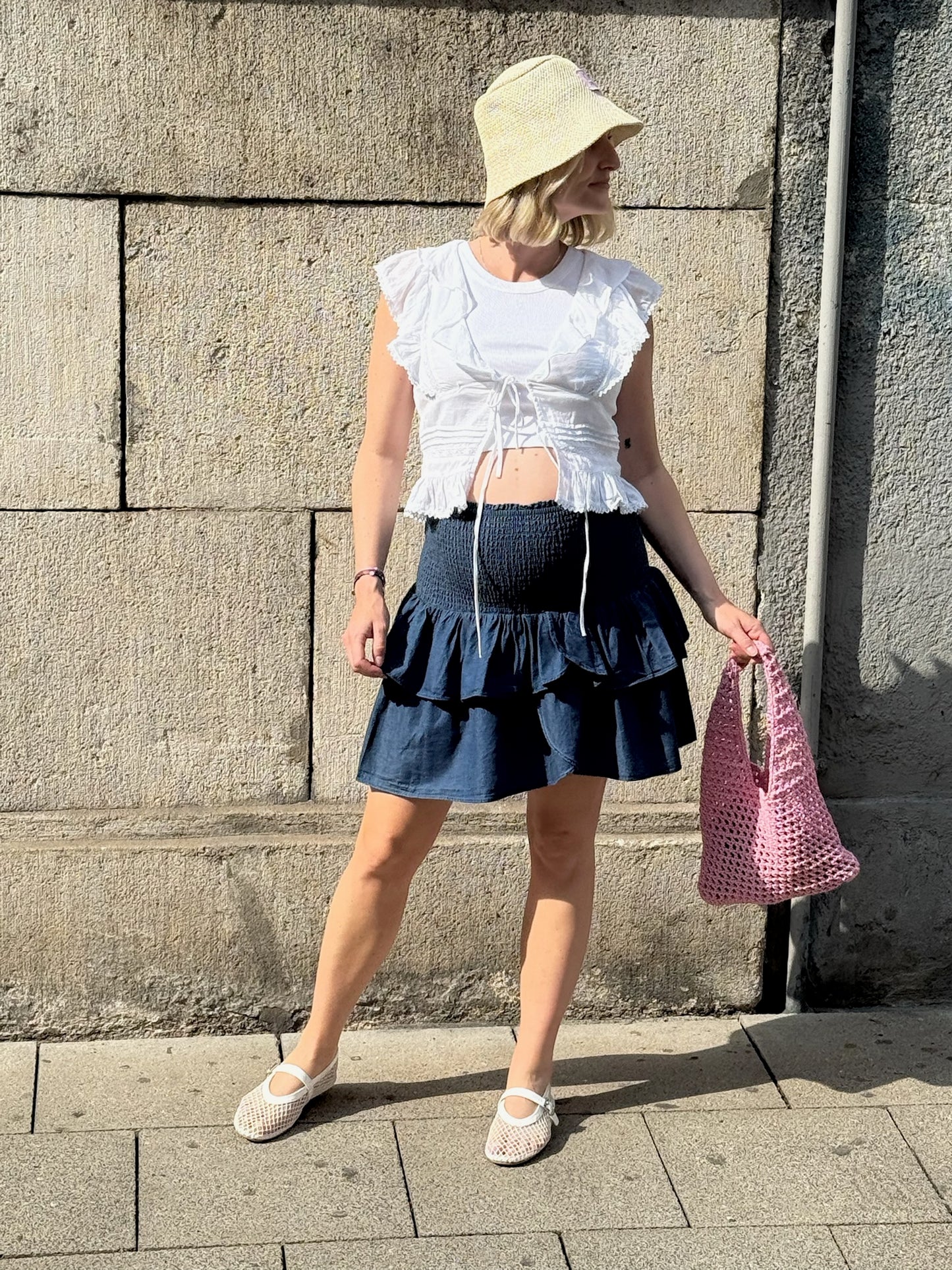 Frau steht vor einer Steinwand in der Sonne. Sie trägt ein weißes, ärmelloses Volant-Top mit Bindebändern über einem sichtbaren Babybauch sowie einen dunkelblauen, kurzen Stufenrock mit gesmoktem Bund. Dazu kombiniert sie einen hellen Sonnenhut, weiße Ballerinas und eine rosa Netztragetasche.