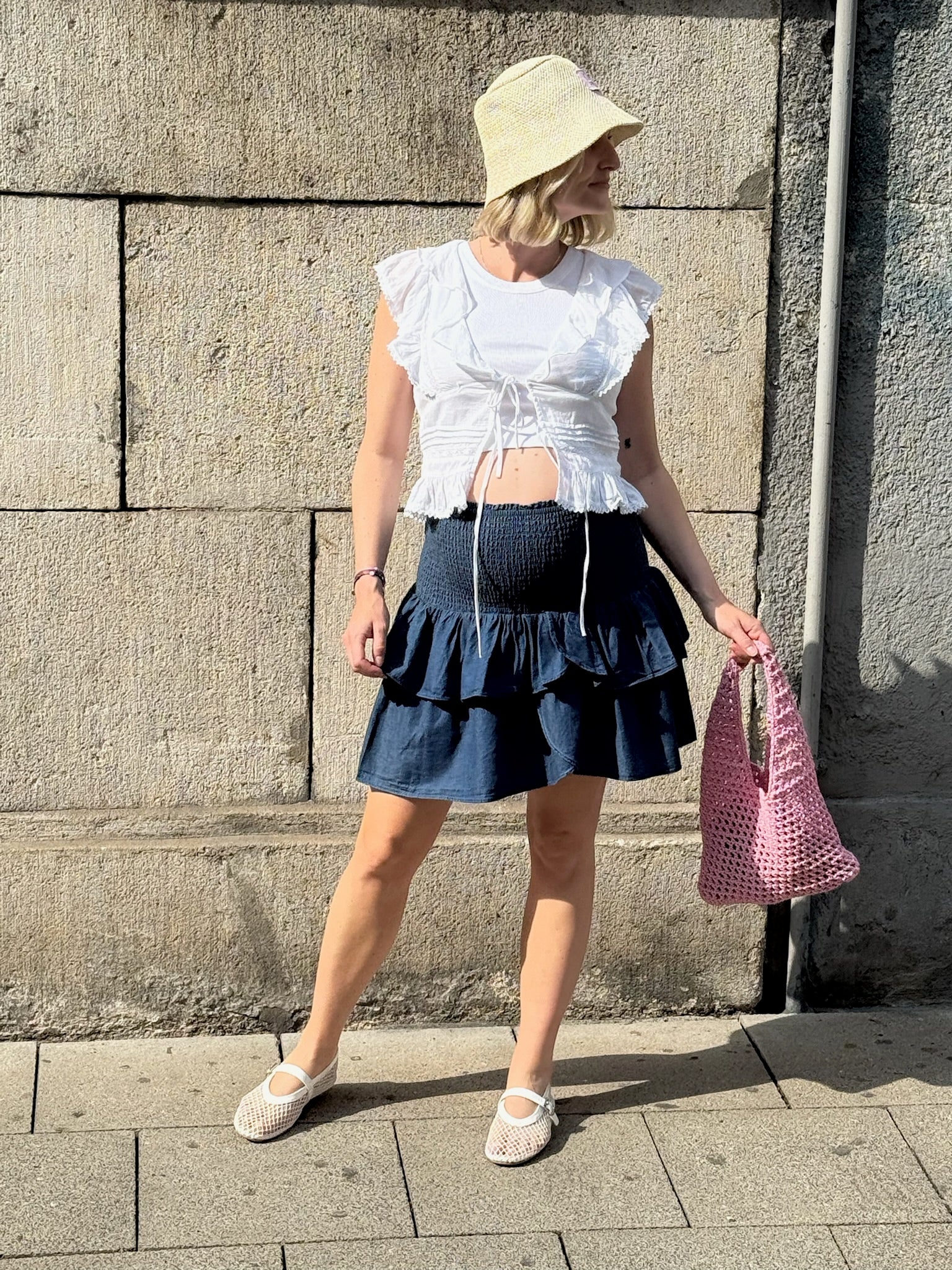 Frau steht vor einer Steinwand in der Sonne. Sie trägt ein weißes, ärmelloses Volant-Top mit Bindebändern über einem sichtbaren Babybauch sowie einen dunkelblauen, kurzen Stufenrock mit gesmoktem Bund. Dazu kombiniert sie einen hellen Sonnenhut, weiße Ballerinas und eine rosa Netztragetasche.