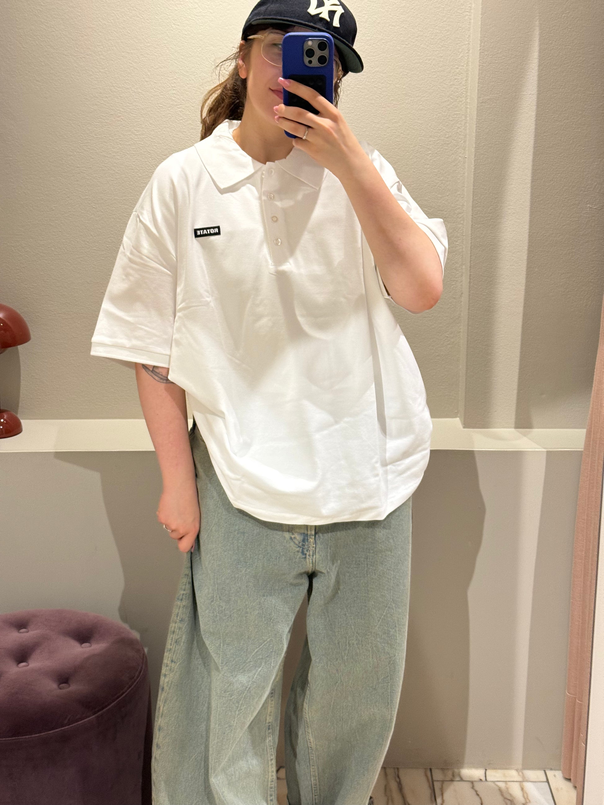Eine Person steht in einer Umkleidekabine und macht ein Spiegel-Selfie. Sie trägt ein weißes, kurzärmeliges Polohemd mit klassischem Kragen und Knopfleiste. Auf der linken Brust befindet sich ein schwarzer Patch mit weißem Schriftzug ‚ROTATE‘.