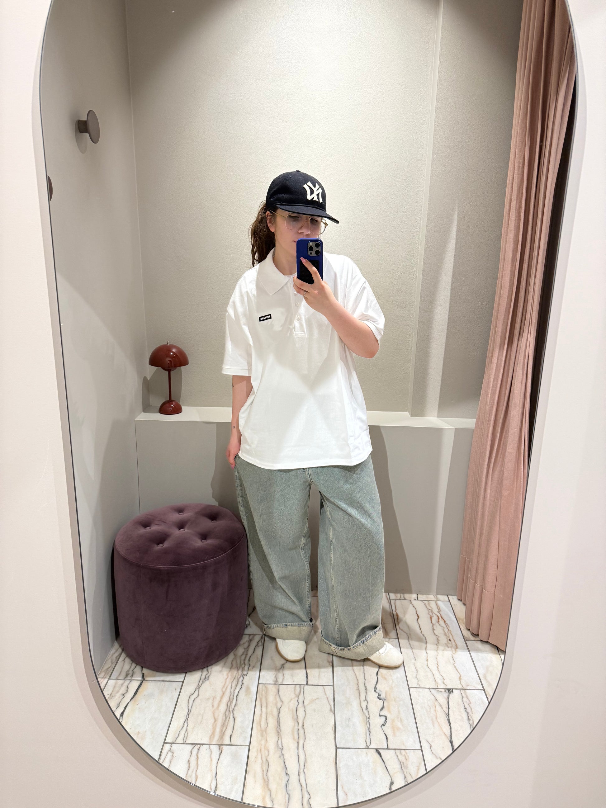 Eine Person steht in einer Umkleidekabine und macht ein Spiegel-Selfie. Sie trägt ein weißes, kurzärmeliges Polohemd mit klassischem Kragen und Knopfleiste. Auf der linken Brust befindet sich ein schwarzer Patch mit weißem Schriftzug ‚ROTATE‘.