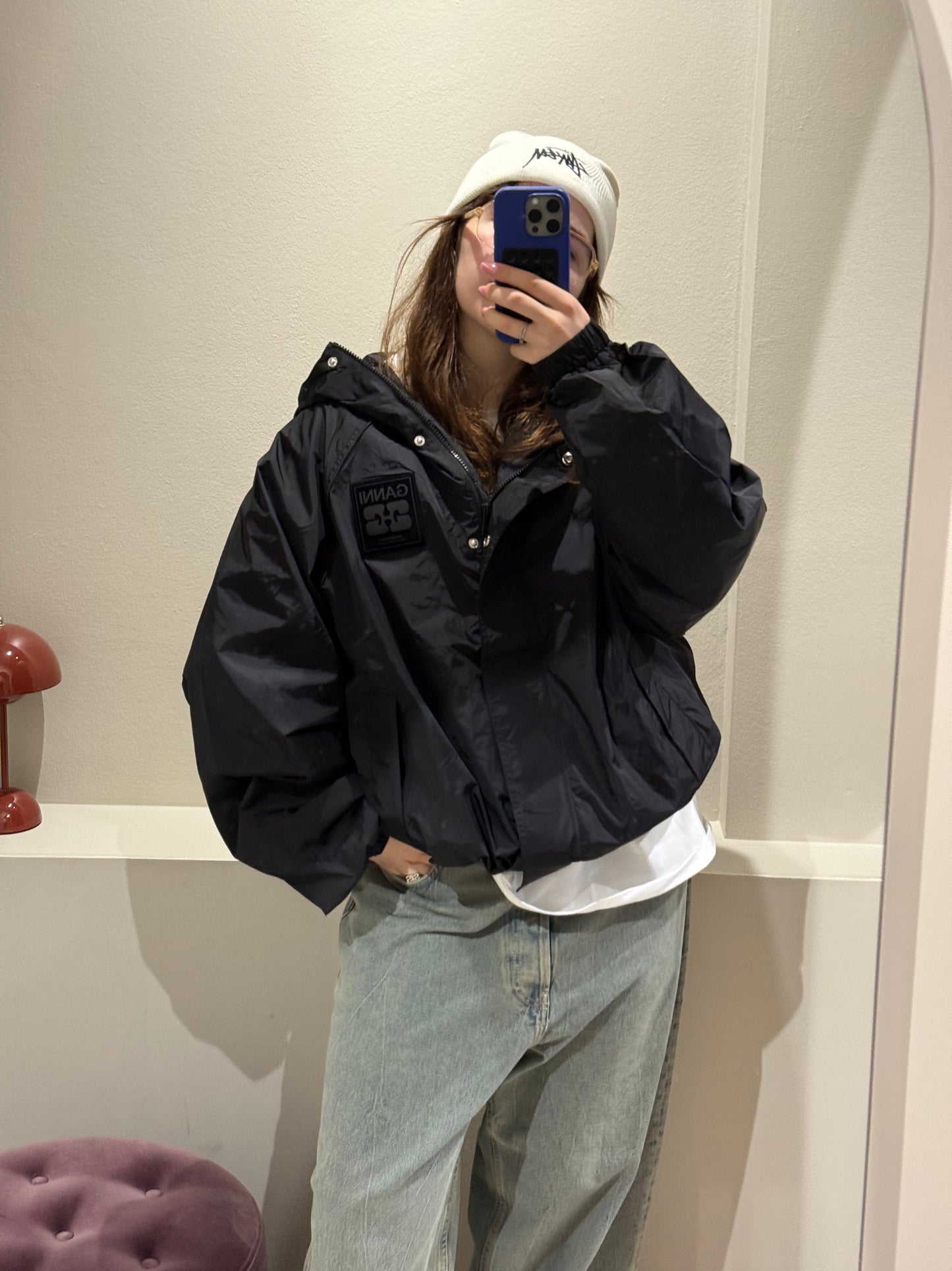 Eine Frau steht in einem Raum vor einem Spiegel und macht ein Selfie. Sie trägt eine schwarze, leicht glänzende Nylonjacke mit Reißverschluss, hohem Kragen und breiten Bündchen an Ärmeln und Saum. Die Jacke hat einen sportlich-kurzen Schnitt.