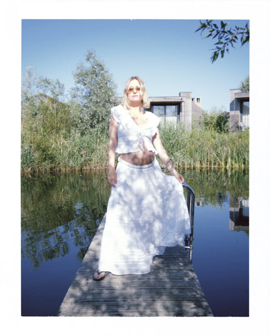 Eine Frau steht auf einem Steg, sie trägt einen weißen fließenden Rock mit horinzontalen Lochstick-Streifen und Rüsschensaum. Dazu trägt sie noch eine Bindebluse mit Voilants und kurzen Ärmle sowie Flip Flops.