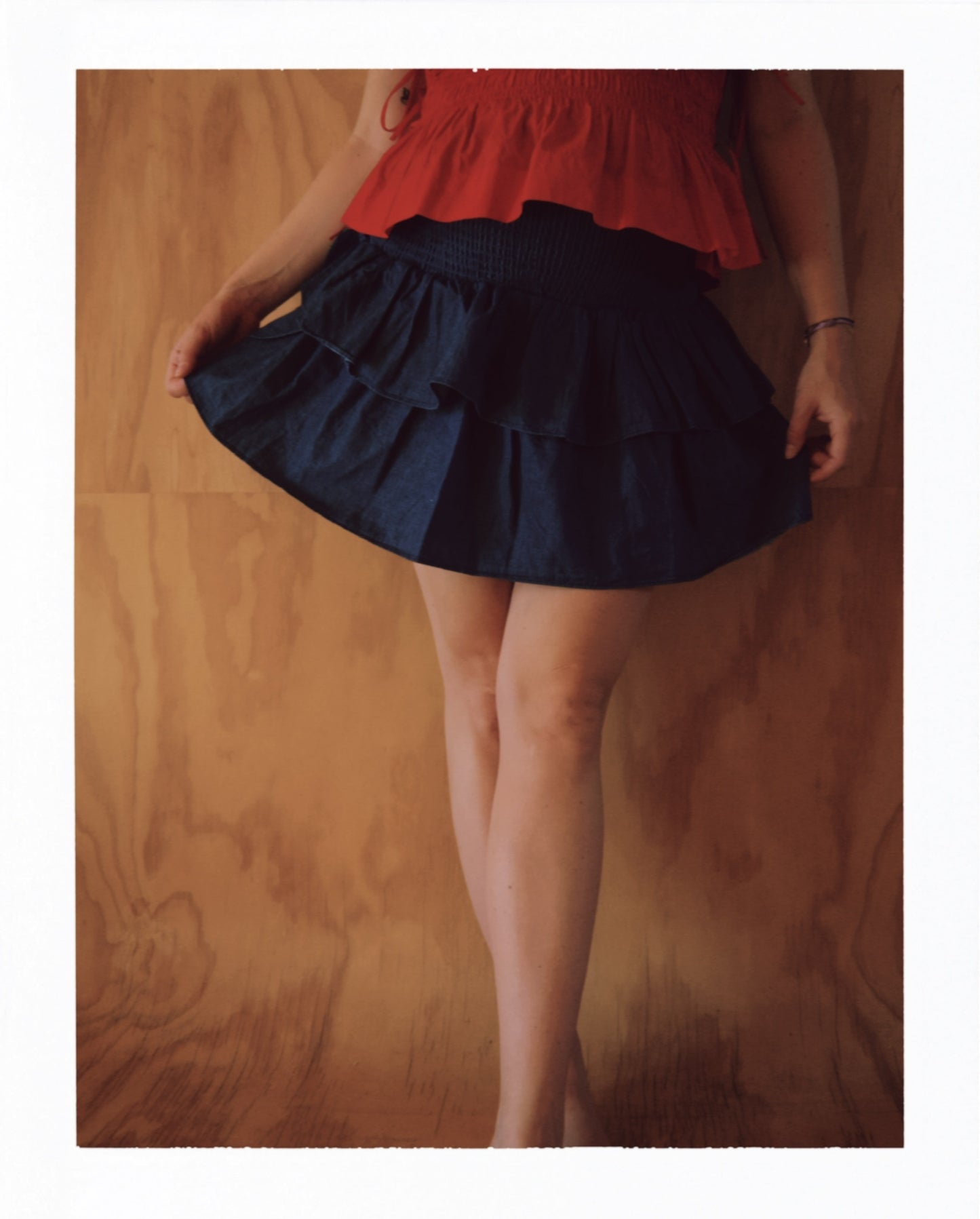 Frau steht barfuß vor einer holzvertäfelten Wand. Sie trägt ein ärmelloses, rotes Oberteil mit Rüschen am Saum und einen kurzen, dunkelblauen Jeansrock mit gesmoktem Bund und doppeltem Volant. Die Pose ist leicht verspielt, mit einem Bein locker vor das andere gestellt.