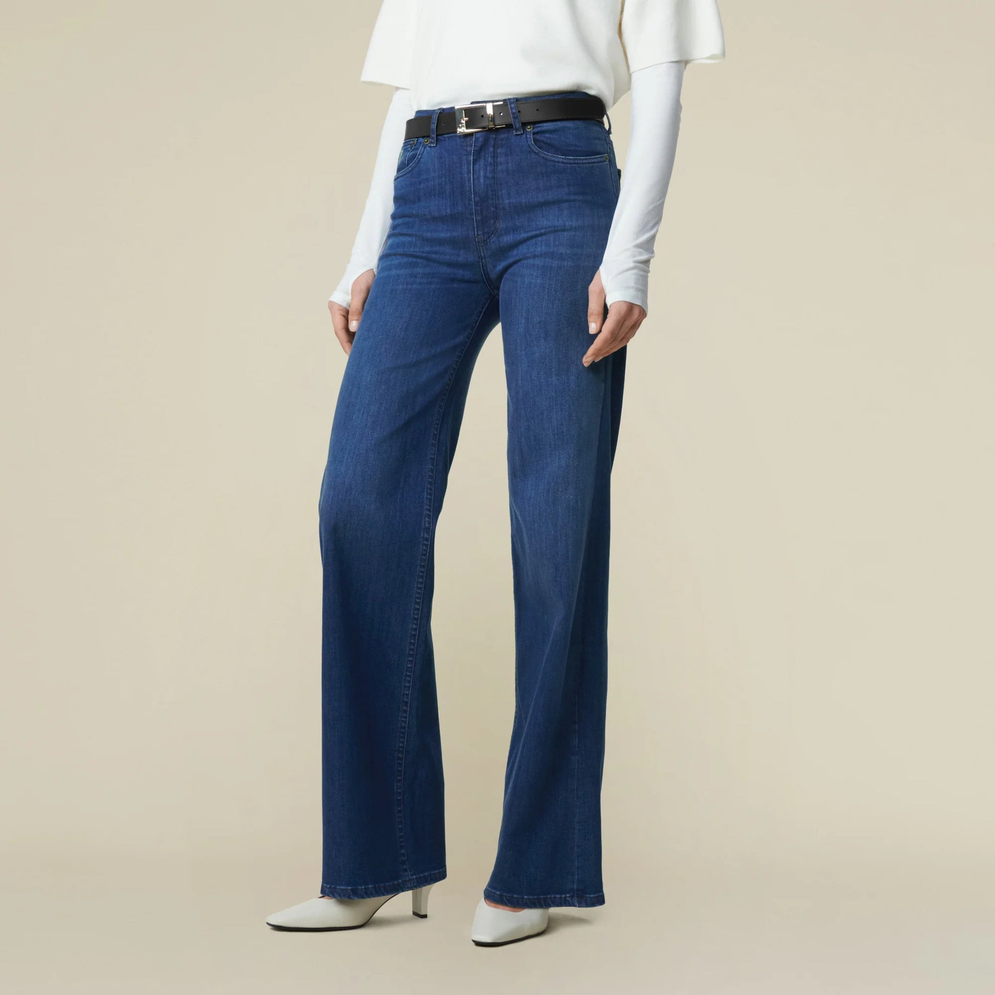 Frau vor weißer Wand, man sieht nur den Unterkörper und eine Teil vom Rumpf. Sie trägt eine mittelblaue Jeans mit weitem Bein und Weiße Schuhe. Zur Jeans Kombiniert sie weißes Oberteil und schwarzen Gürtel. 