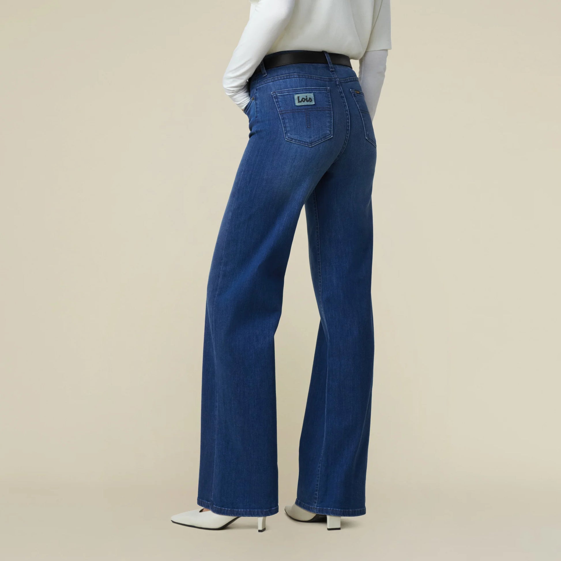 Frau vor weißer Wand, man sieht nur den Unterkörper und eine Teil vom Rumpf. Sie trägt eine mittelblaue Jeans mit weitem Bein und Weiße Schuhe. Zur Jeans Kombiniert sie weißes Oberteil und schwarzen Gürtel. Man sieht sie von hinten.