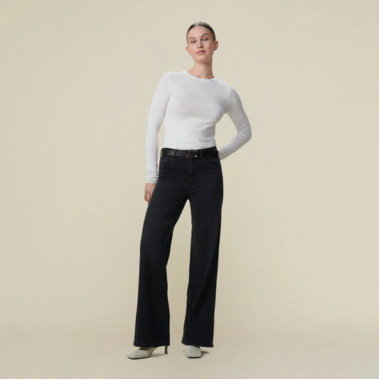 rau vor weißer Wand, man sieht nur den Unterkörper und eine Teil vom Rumpf. Sie trägt eine schwarze Jeans mit weitem Bein und Weiße Schuhe. Zur Jeans Kombiniert sie weißes Oberteil und schwarzen Gürtel. 