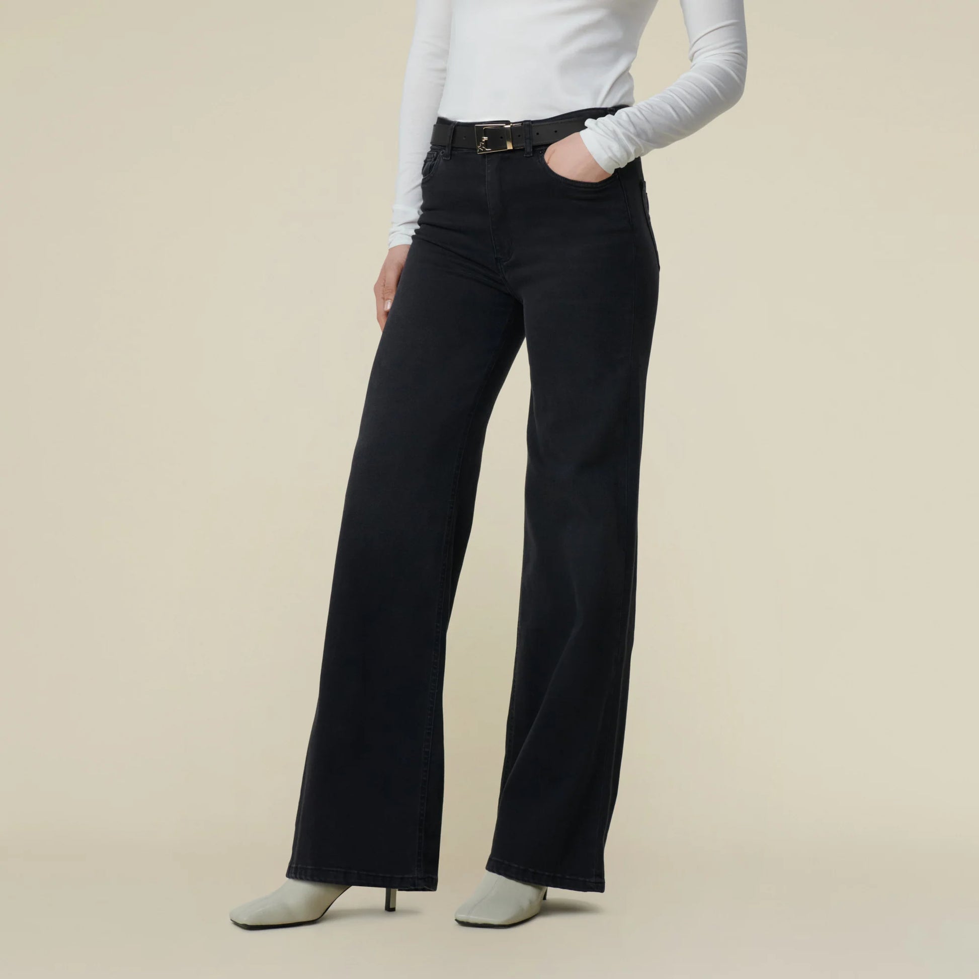 rau vor weißer Wand, man sieht nur den Unterkörper und eine Teil vom Rumpf. Sie trägt eine schwarze Jeans mit weitem Bein und Weiße Schuhe. Zur Jeans Kombiniert sie weißes Oberteil und schwarzen Gürtel. 