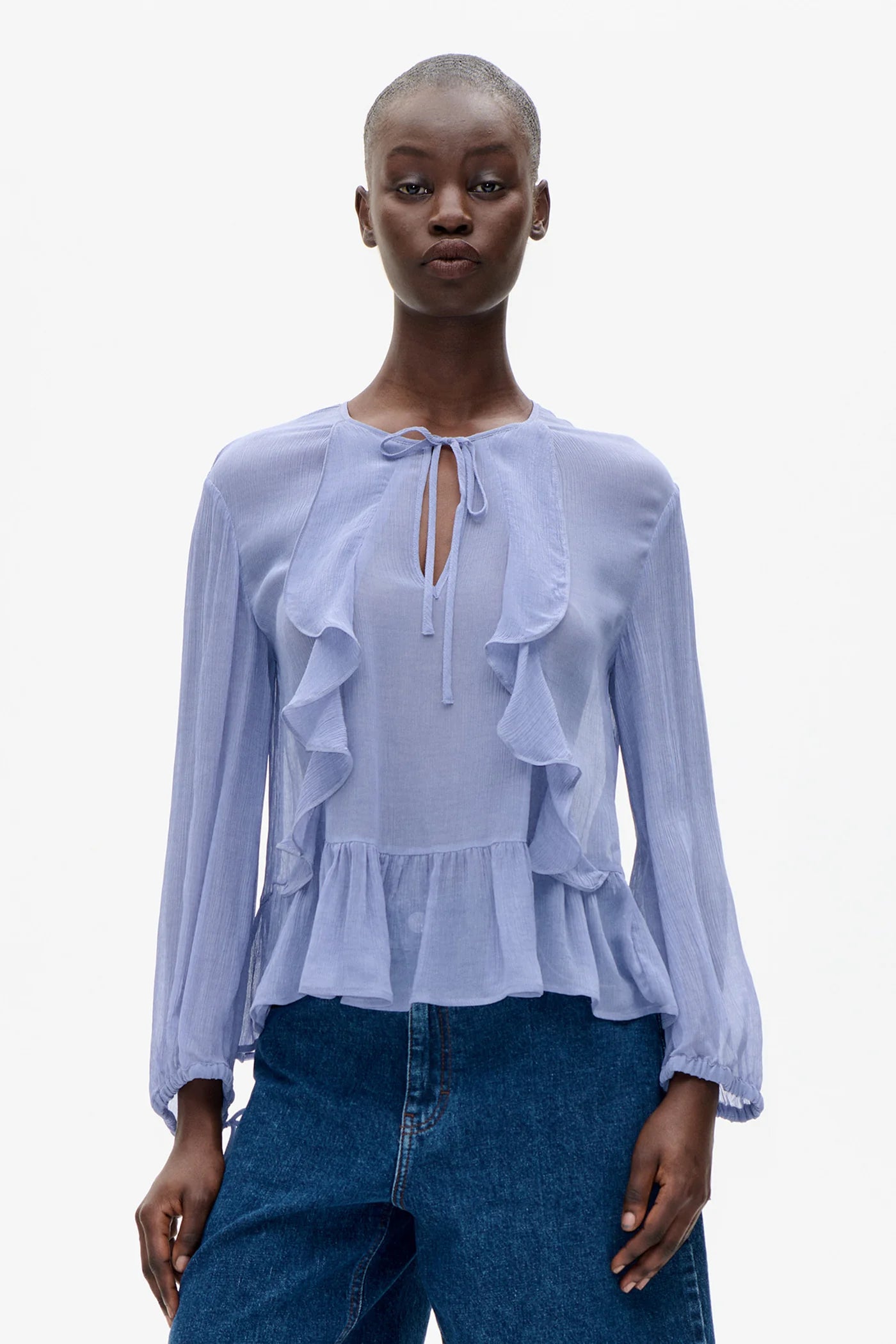 Eine Person steht frontal vor hellem Hintergrund und trägt eine puderblaue Bluse mit Volants, Bindekragen und langen Ärmeln. Die Bluse ist leicht transparent und fällt locker über eine blaue Jeans.