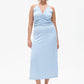 Frau trägt ein hellblaues, ärmelloses Midikleid mit Spaghettiträgern und V-Ausschnitt. Das Kleid hat eine geraffte Partie im Brustbereich und fällt figurbetont bis midiwadenlang. Sie kombiniert es mit schwarzen Slingback-Pumps mit kleinem Absatz. Der Hintergrund ist weiß und neutral gehalten.