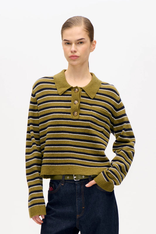 Frau vor weißer Wand, sie trägt eine dunkle Jeans und ein grünes Polo mit schwarzen und weißesn streifen. Das Polo hat eine Brusttasche und braune Knöpfe.
