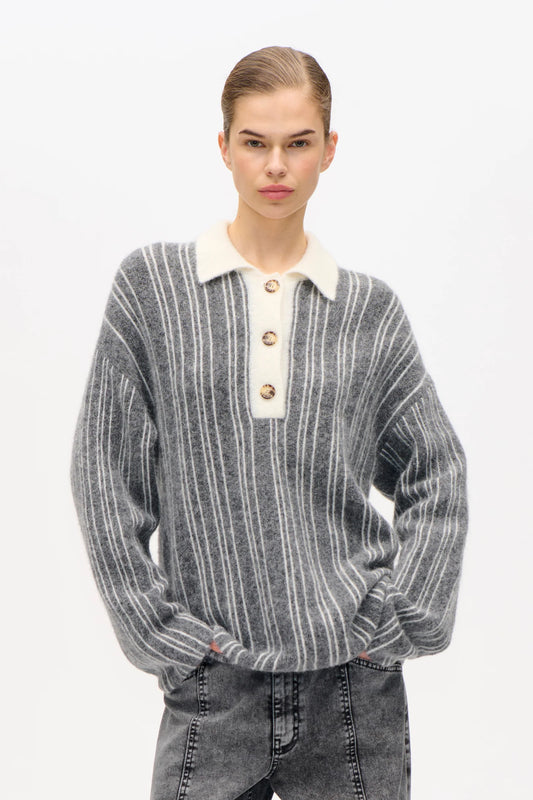Frau vor weißer Wand, sie trägt Graue Jeans und ein großes Oversized Strickpolo in Grau mit vertikalen Streifen und farblich abgesetzten Cremfarbenens Kragen und Knopfleiste.