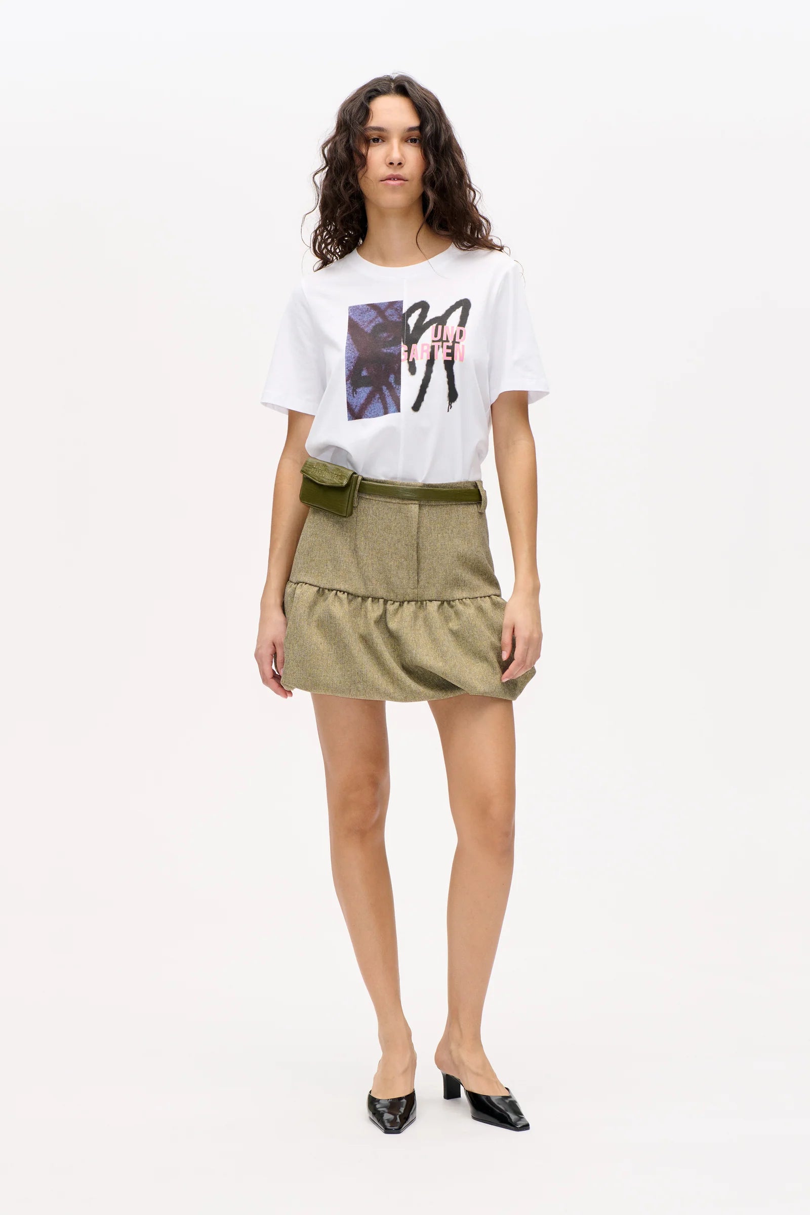 Frau vor weißer Wand, sie trägt einen grünen Tweed-Rock und weißes Printshirt.