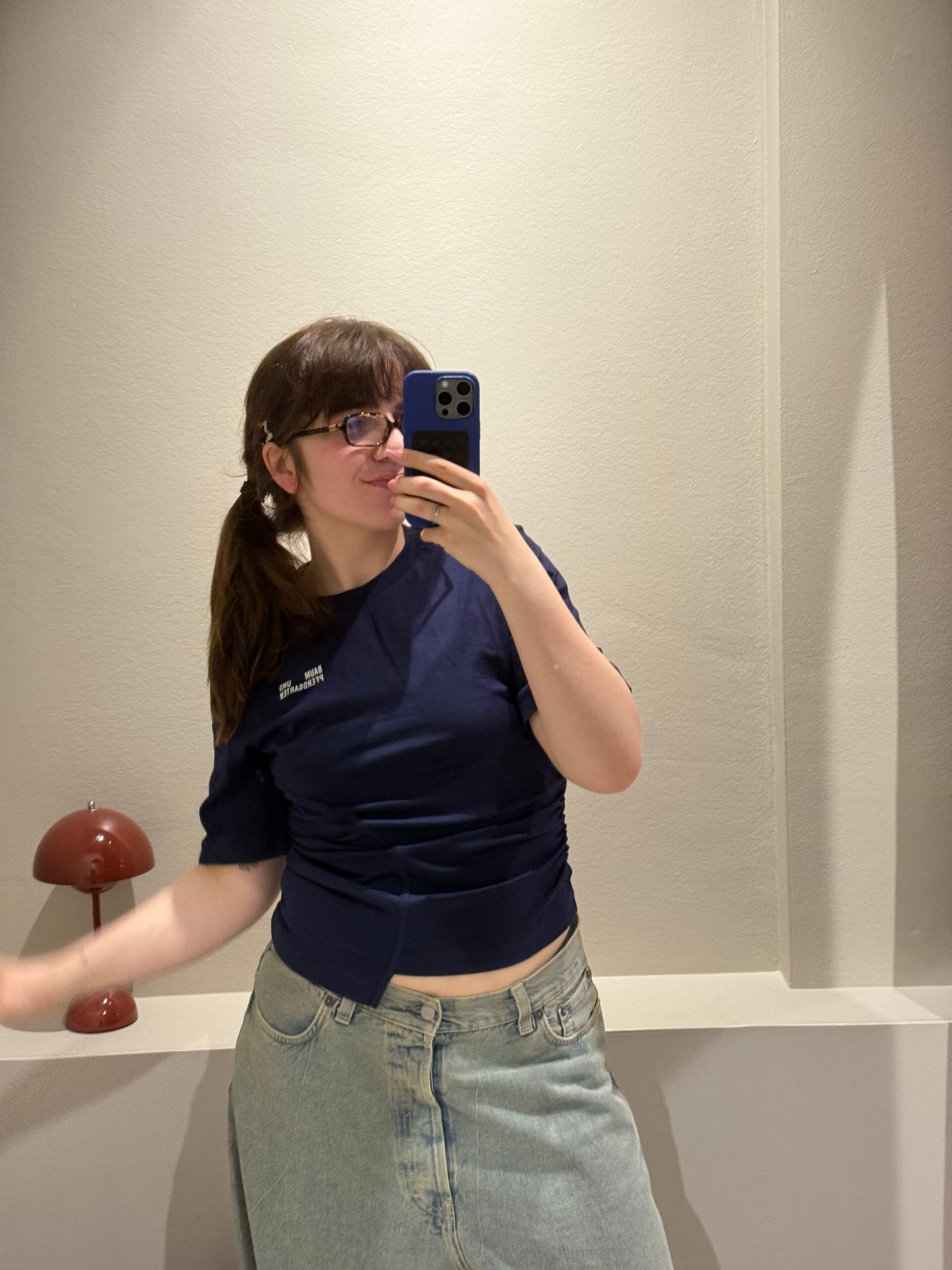 Frau trägt ein dunkelblaues, tailliertes T-Shirt mit asymmetrischem Schnitt und seitlicher Raffung. Auf der linken Brust befindet sich eine kleine Logo-Aufschrift ‚Baum und Pferdgarten‘. Dazu kombiniert sie eine hellblaue Jeans mit weitem Bein. Sie steht in einer Umkleidekabine mit hellem Vorhang und violettem Hocker.