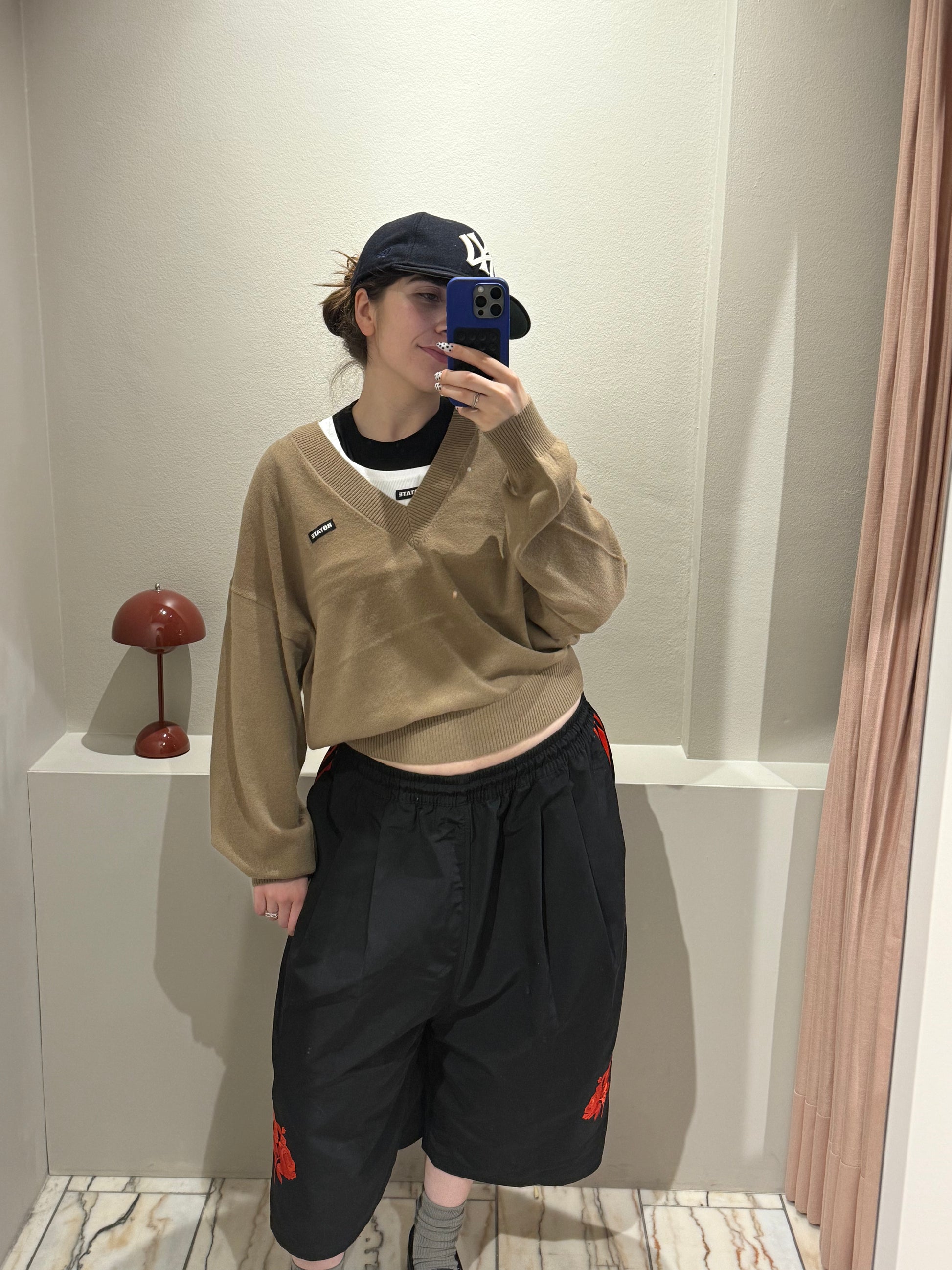 Frau macht Spiegelselfie, sie trägt eine schwarze Hose und dazu einen camelfarbenen V-Neck Strick mit ROTATE Logopatch auf der rechten Brust.