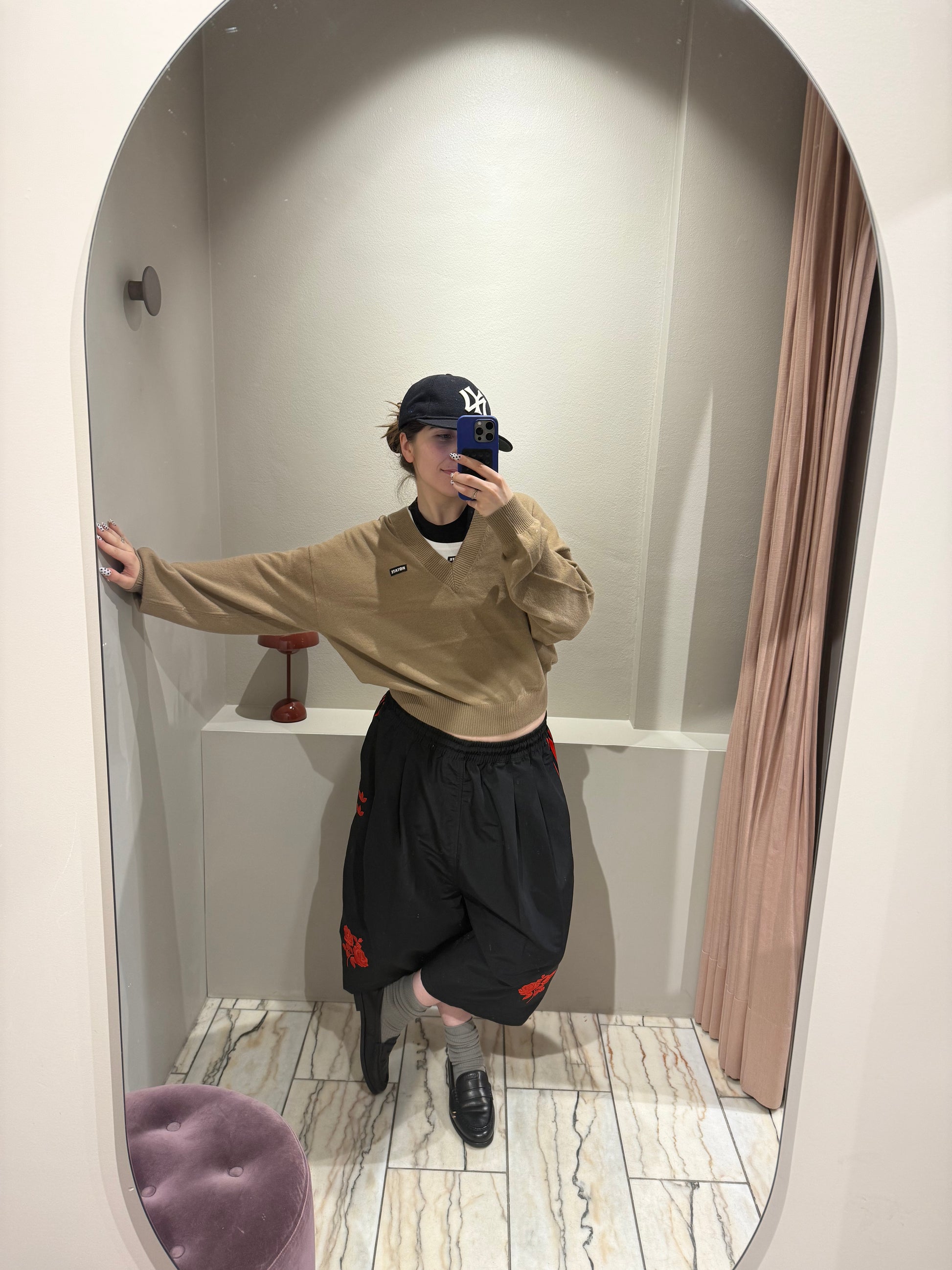 Frau macht Spiegelselfie, sie trägt eine schwarze Hose und dazu einen camelfarbenen V-Neck Strick mit ROTATE Logopatch auf der rechten Brust.
