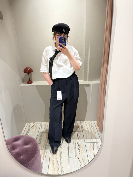 Frau macht Spiegelselfie, sie trägt eine dunkelblaue Bundfaltenhose mit weitem Bein. Dazu ein weißes Polo mit schwarzer Krawatte und Loafer.