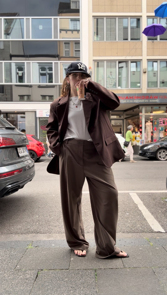 Frau trinkt Kaffe, sie trägt eine braune Hose mit einem overiszed Blazer, der ist schokobraun. Im Hintergrund sieht man eine Straße.
