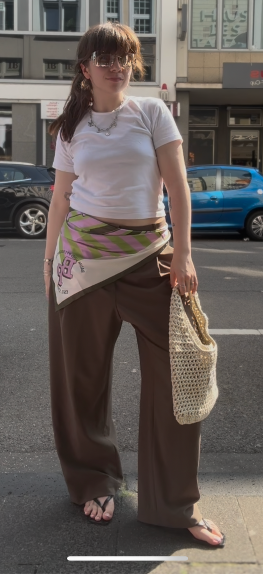 Frau auf der Straße, sie trägt eine Weite Bundfaltenhose mit weißem Tshirt und Flip Flops. Um die Hose hat sie noch ein Seidentuch in grün und Pinktönen, dazu hat sie noch eine gehäklete Tasche.