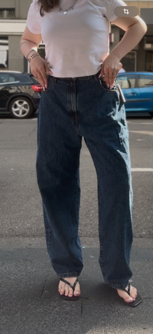 Frau auf der Straße. Sie trägt ein weißes Tshirt mit einer blauen Jeans mit O-Shape, dazu Flip FLops und Silberschmuck.