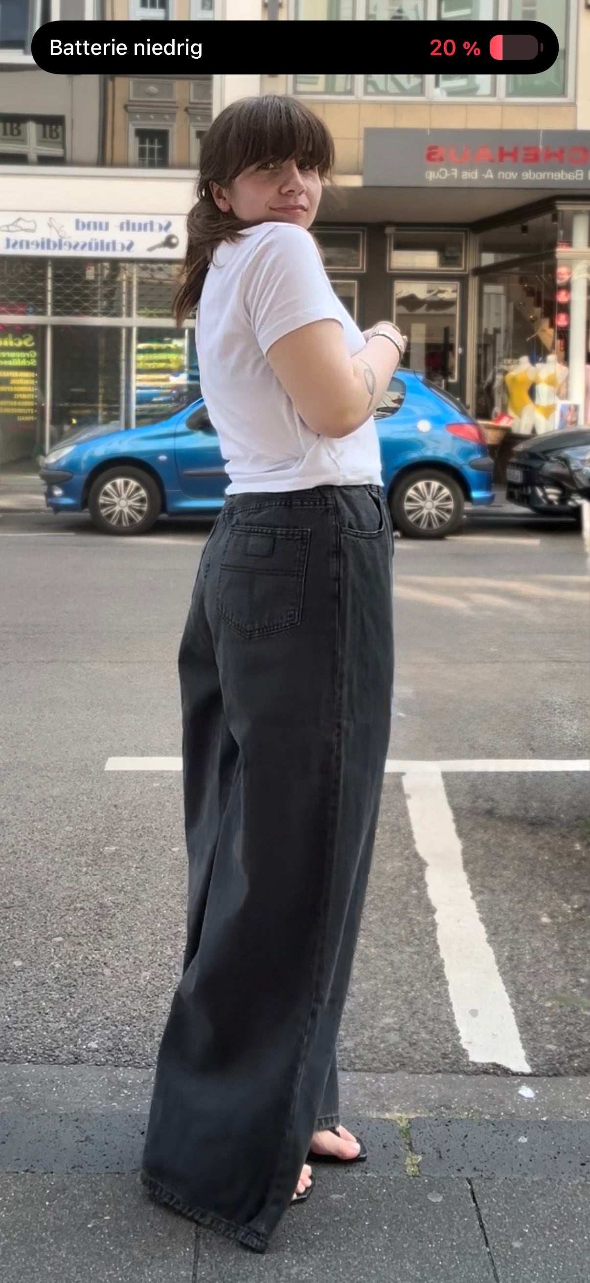 Frau auf der Straße im Seitenprofil. Sie trägt ein weißes Tshirt mit einer extra weitem Jeans in Grau. dazu Flip FLops und Silberschmuck.