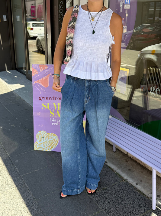 Outfitbild von einer Frau, sie trägt eine gewaschene Jeans mit Bundfalten und ein weißes gesmocktes Top. Dazu Flip Flops und eine Häkeltasche.