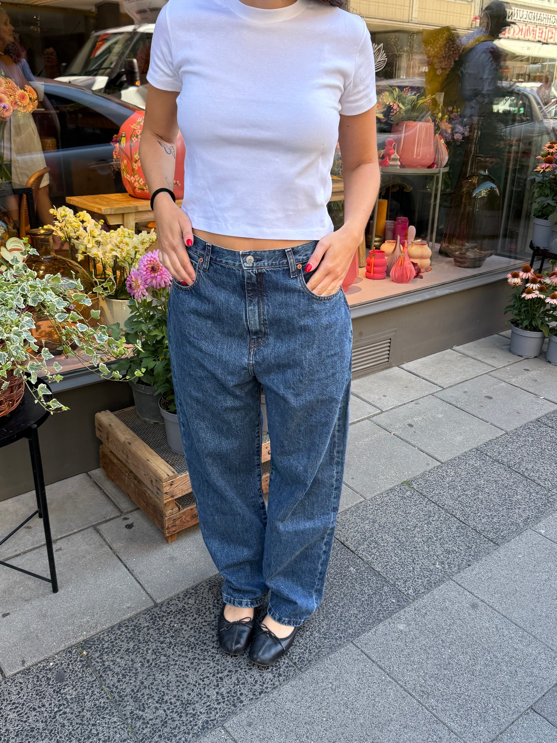 Frau steht vor Blumenladen, sie trägt eine dunkle Jeans mit Bllerina und einem weißem Tshirt. Die Jeans ist ballonförmig geschnitten.