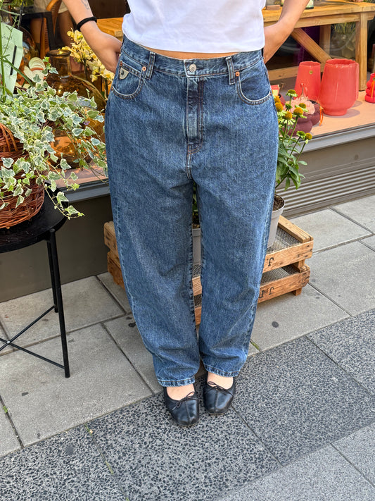 Frau steht vor Blumenladen, sie trägt eine dunkle Jeans mit Bllerina und einem weißem Tshirt. Die Jeans ist ballonförmig geschnitten.