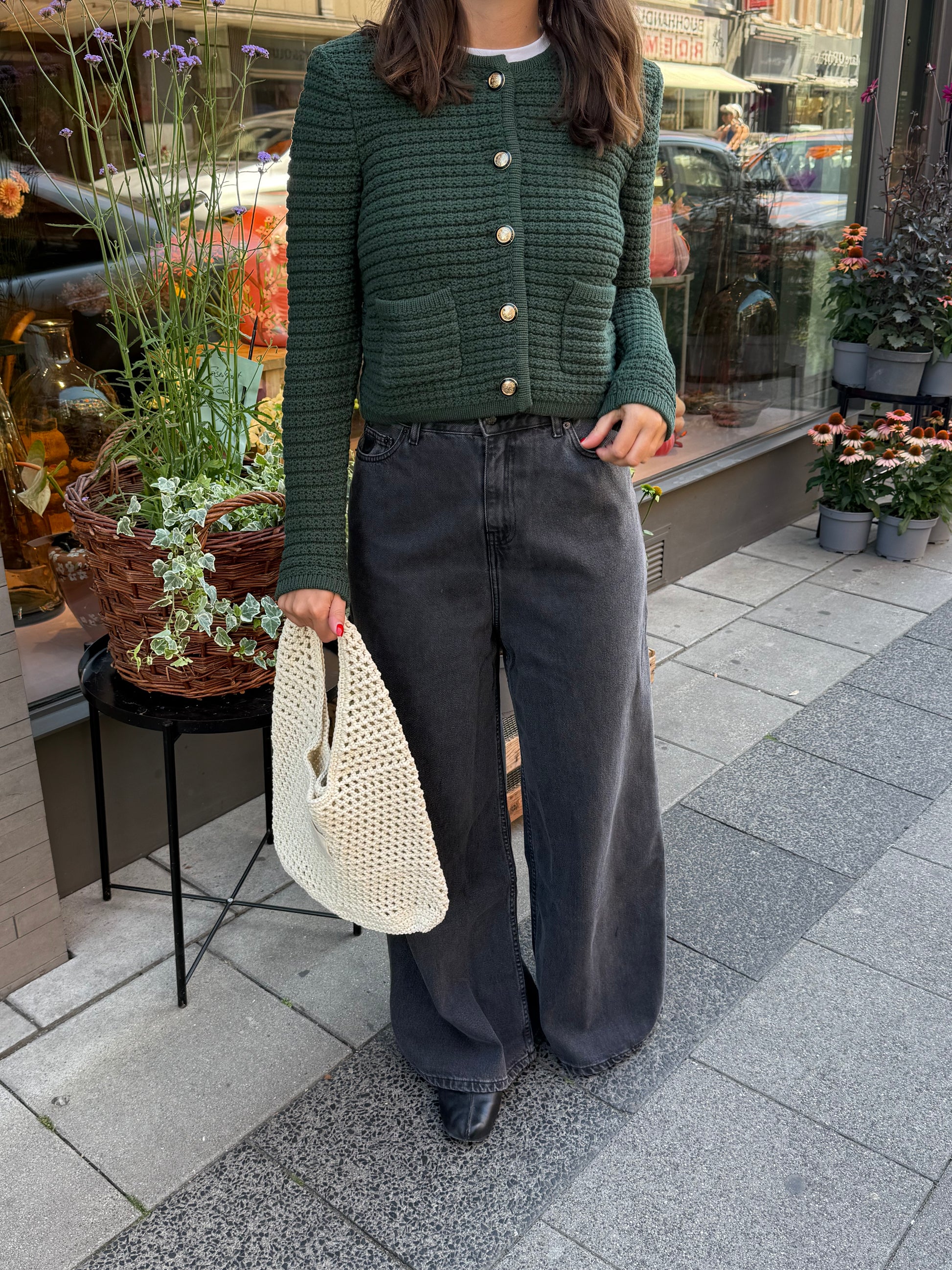 Frau steht vor Blumenladen, sie trägt eine graue weite Denim und einen waldgrünen ardigan mit goldenen Knöpfen. 