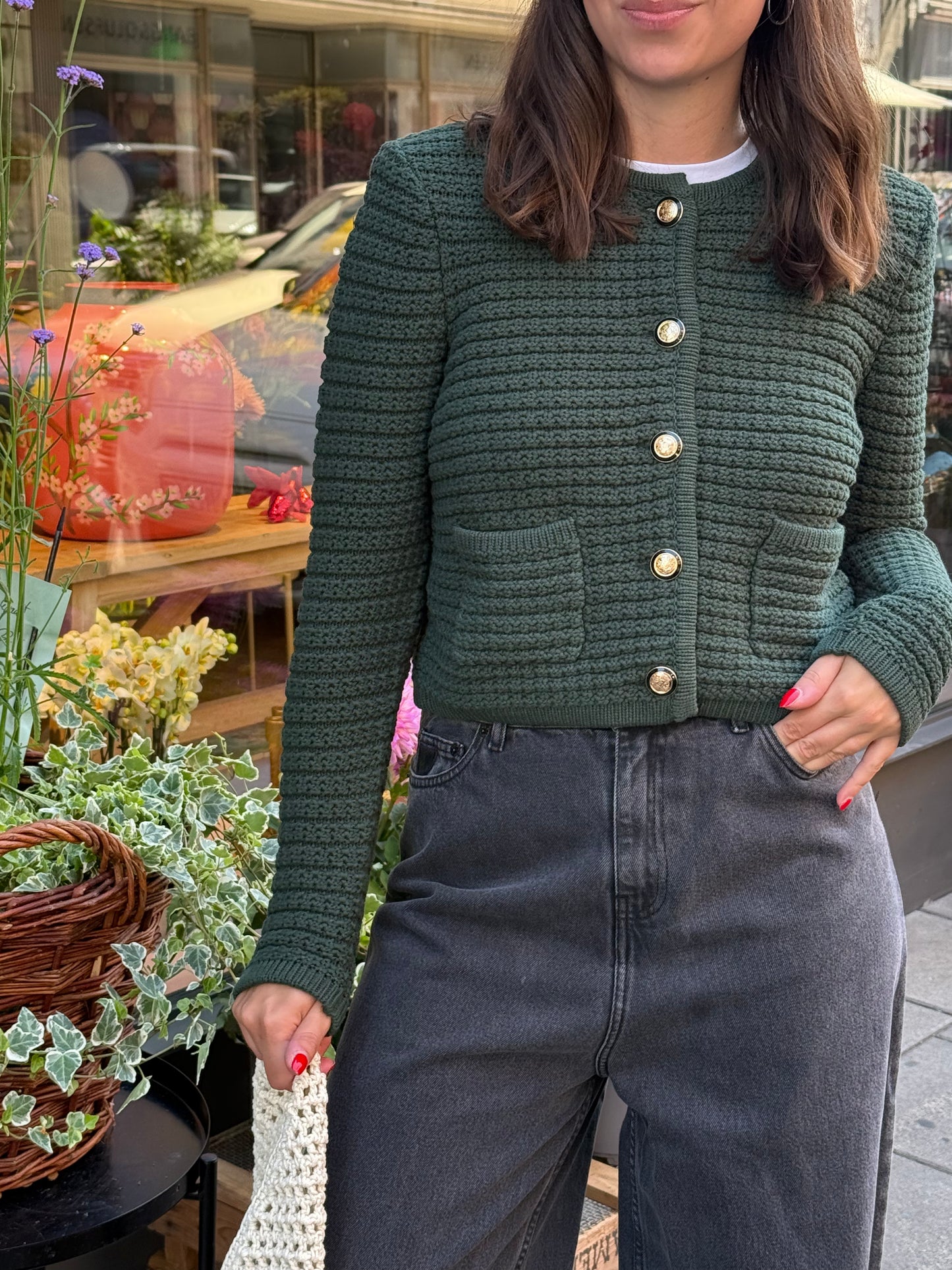 Frau steht vor Blumenladen, sie trägt eine graue weite Denim und einen waldgrünen ardigan mit goldenen Knöpfen. 